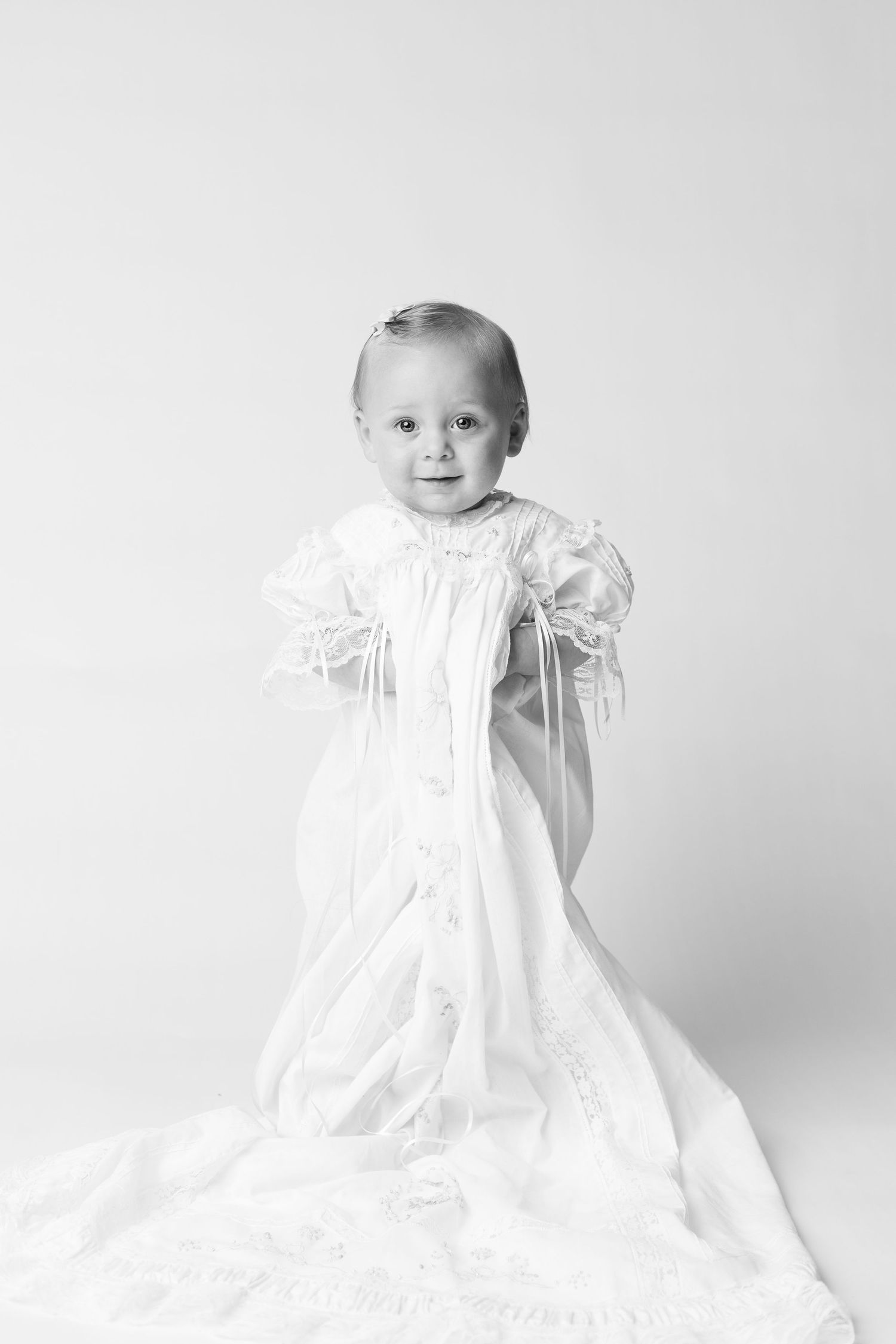 Black and white photo of a smiling baby wearing an elegant white christening gown with lace trim and ribbon details.