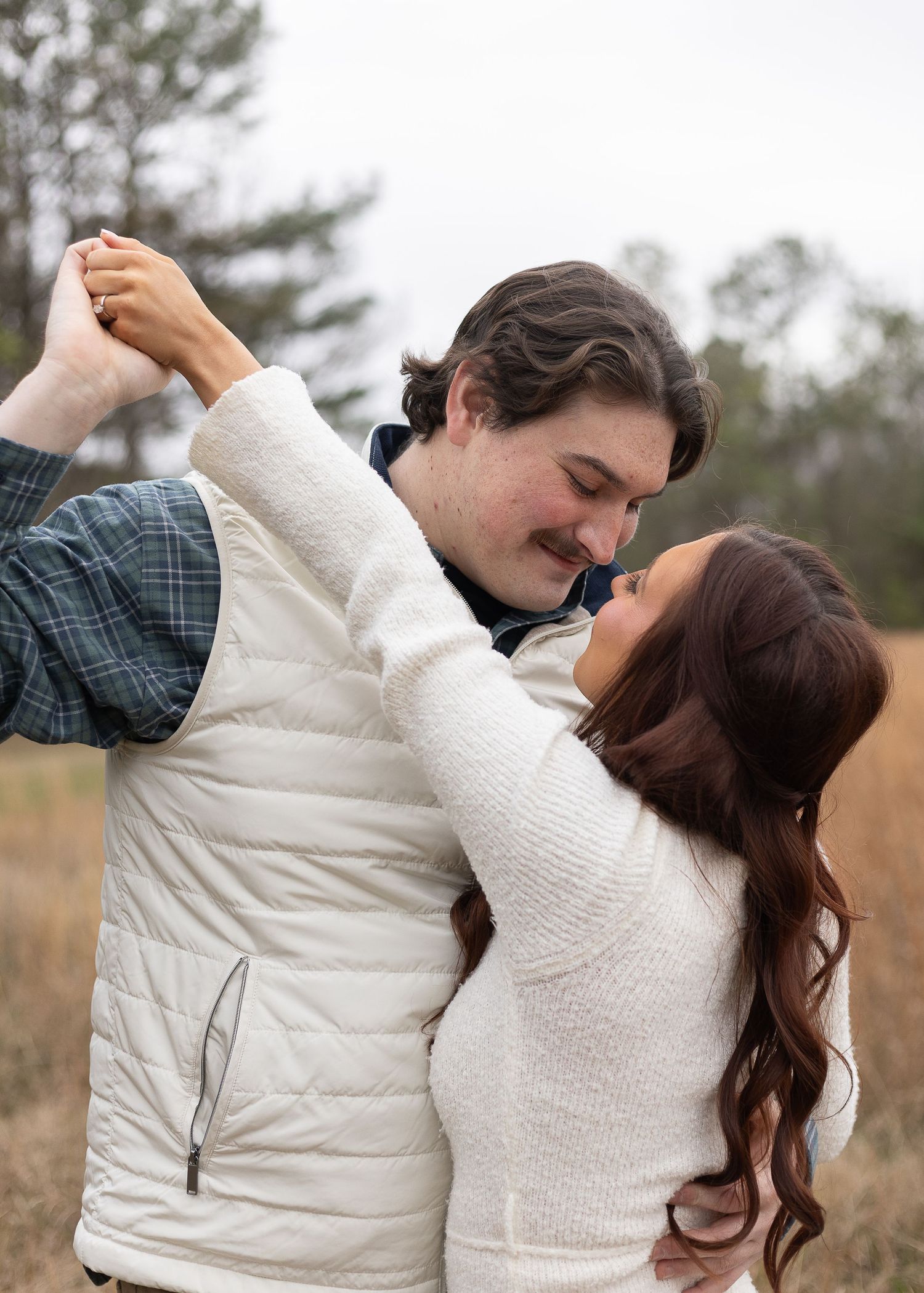 Couple dancing romantically in a field, man in white vest and plaid shirt, woman in cream sweater, overcast sky.