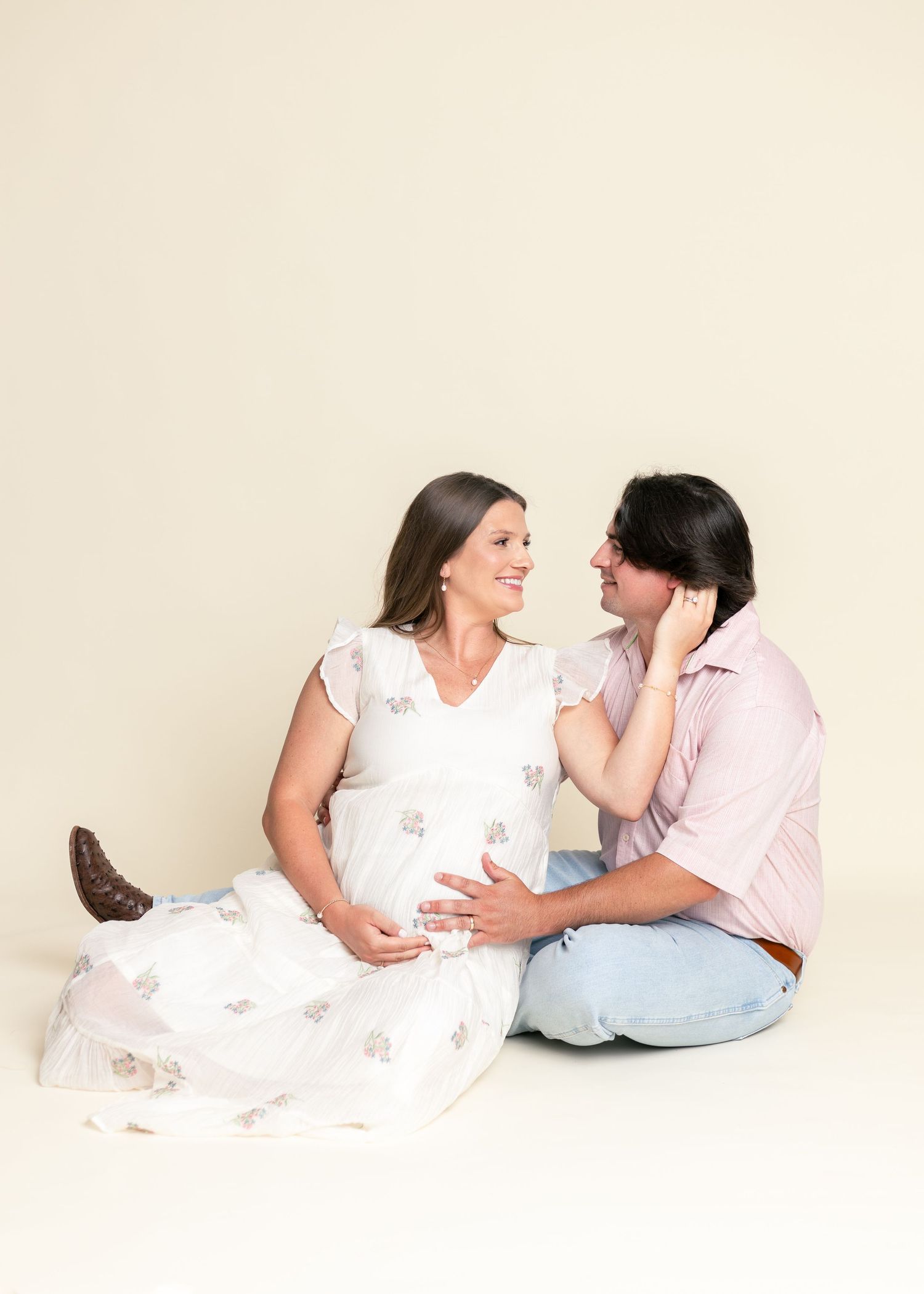 Pregnant woman in white floral dress sits with partner on cream background, sharing a tender smile while cradling her baby bump.