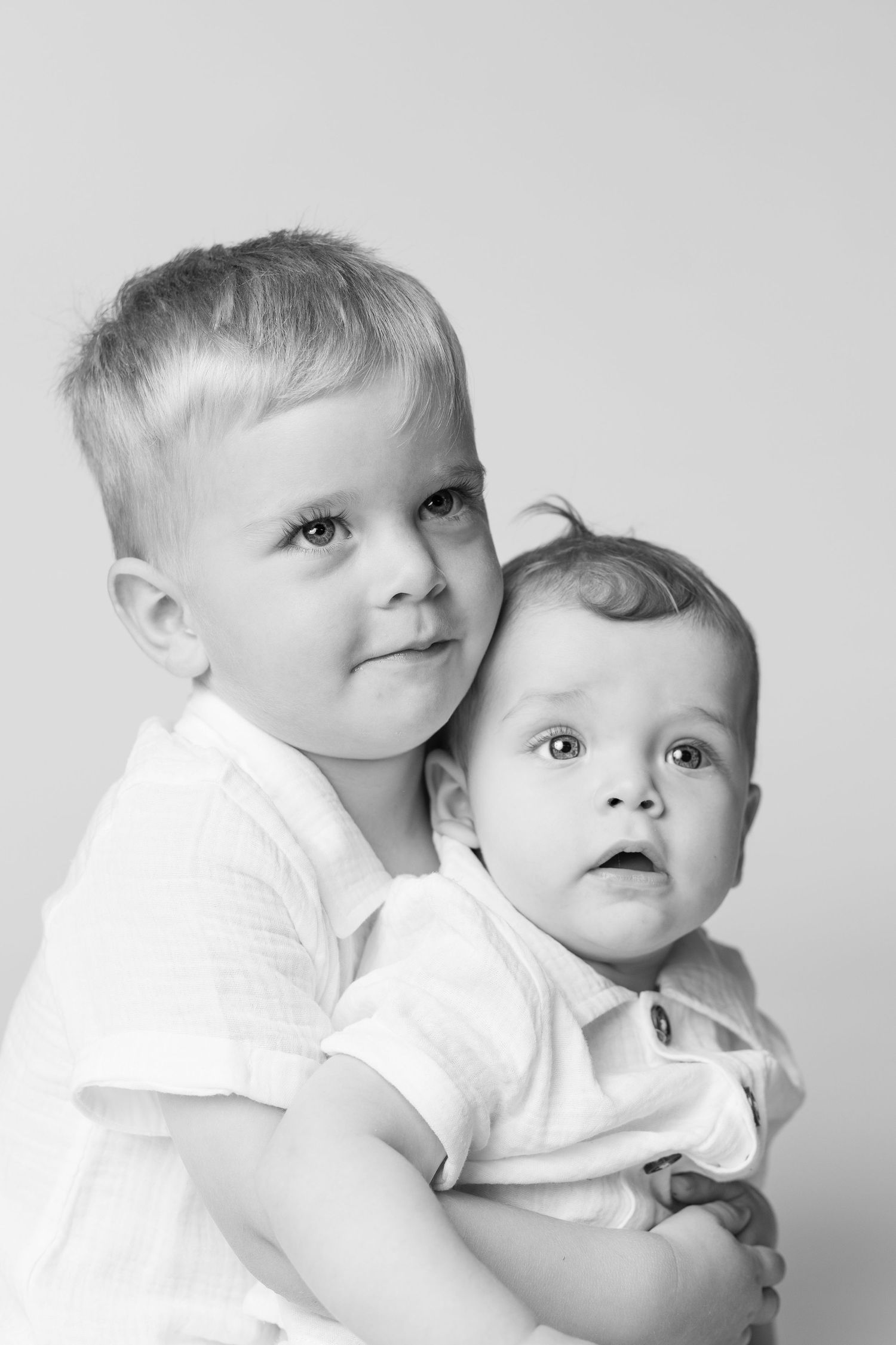 Black and white portrait of an older boy tenderly holding a baby sibling, both wearing white shirts.