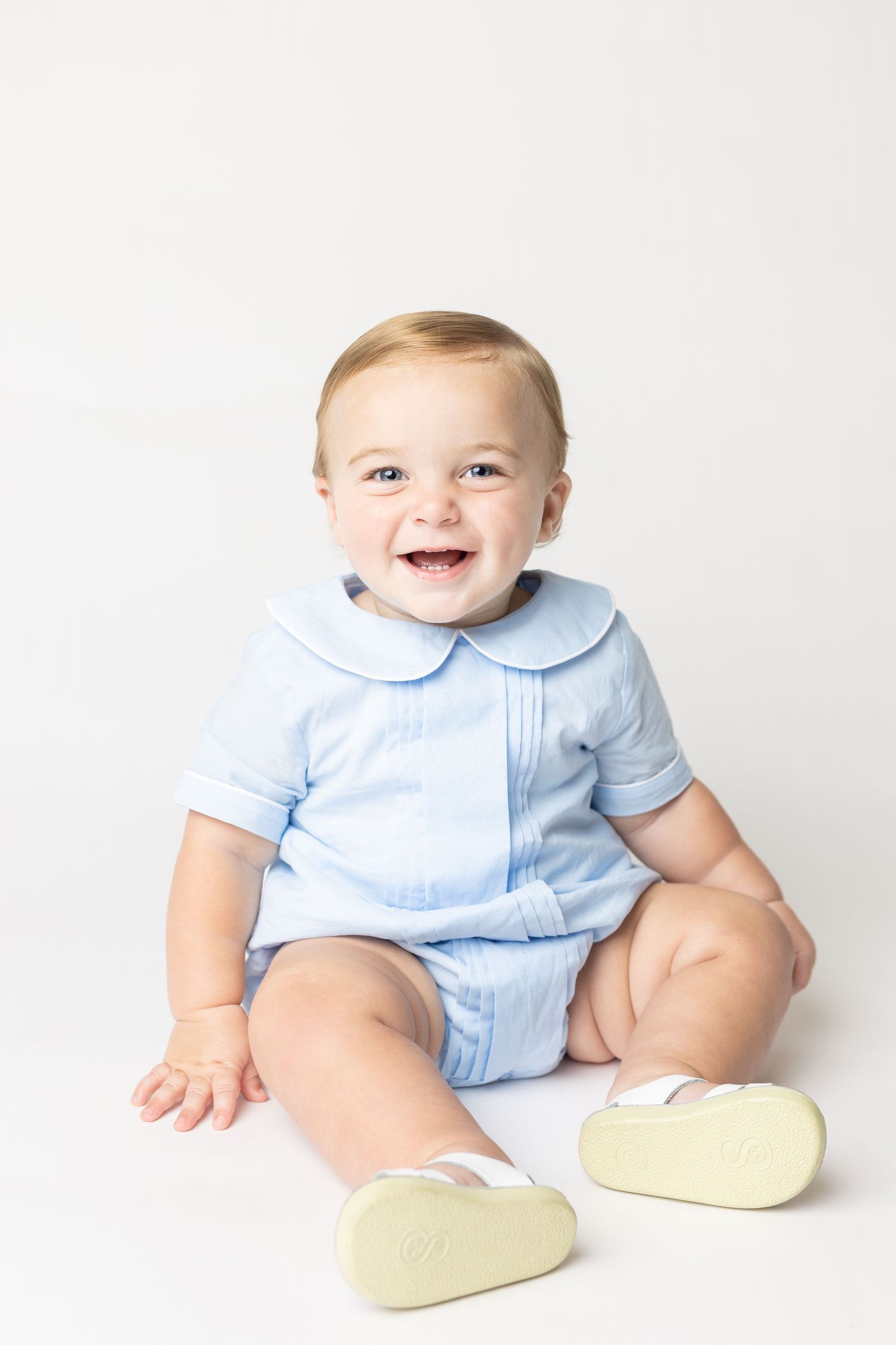 Smiling baby in blue outfit and white shoes sitting on white background.