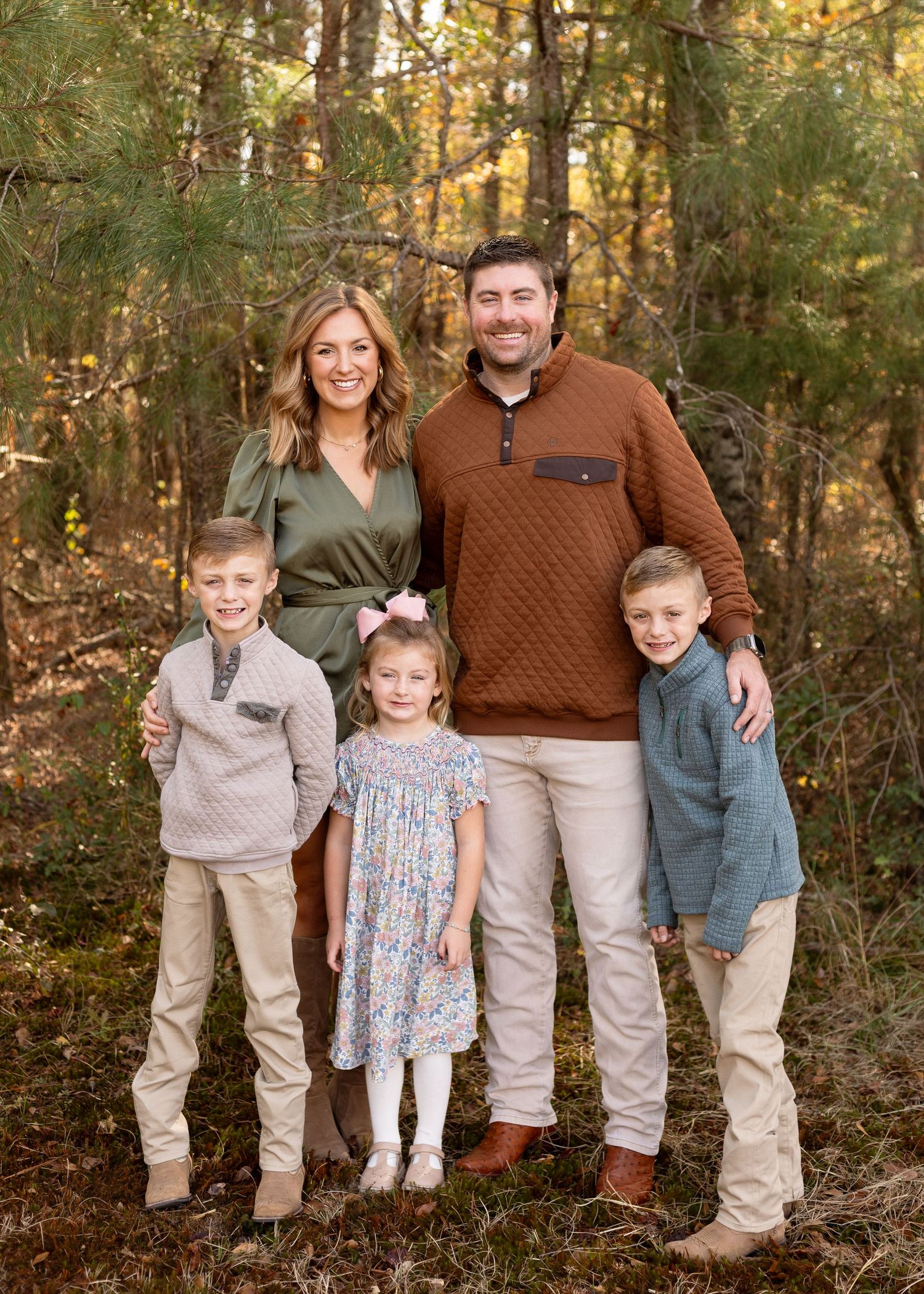 Family of five posing outdoors in coordinated fall outfits among pine trees.