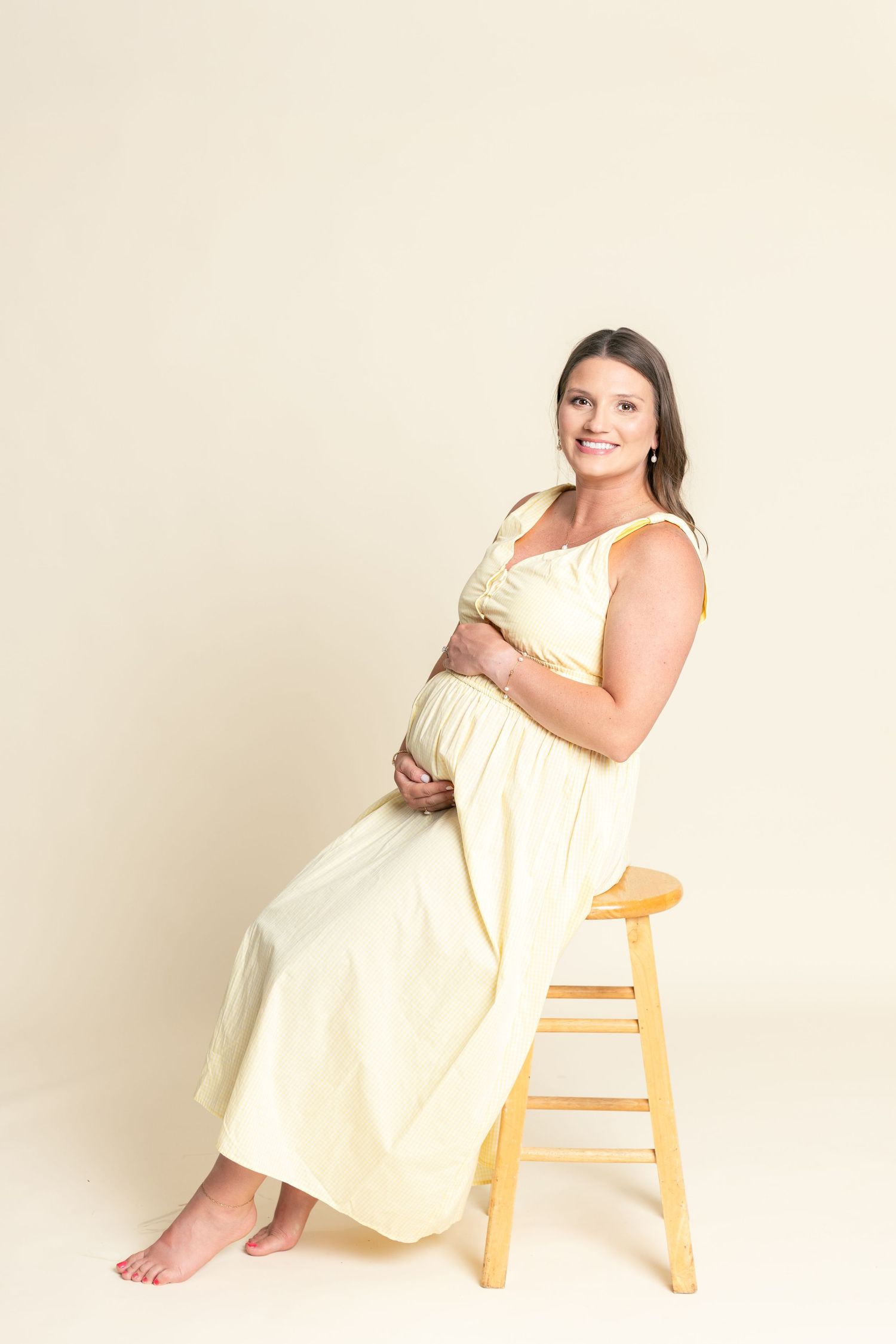 Smiling pregnant woman in yellow dress sitting on wooden stool, cradling baby bump in studio portrait.