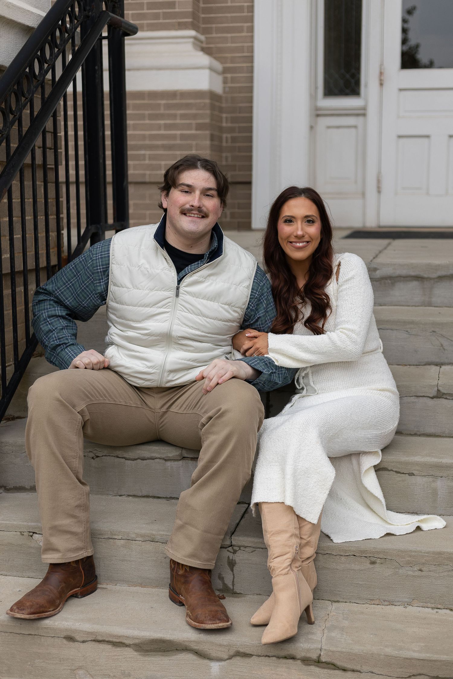A smiling couple sits together on stone steps, man in vest and khakis, woman in white dress and boots.