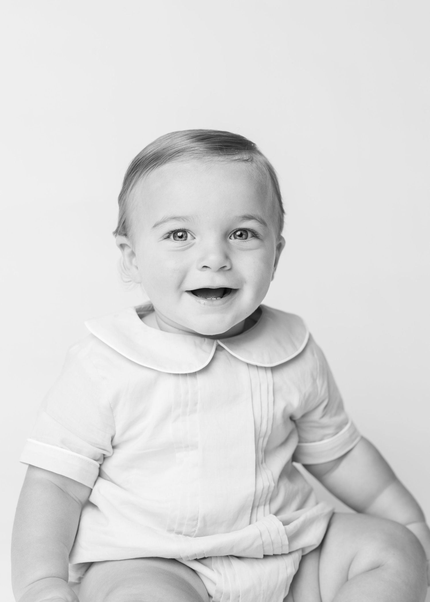 Black and white portrait of a smiling baby in a white Peter Pan collar outfit, studio photography.