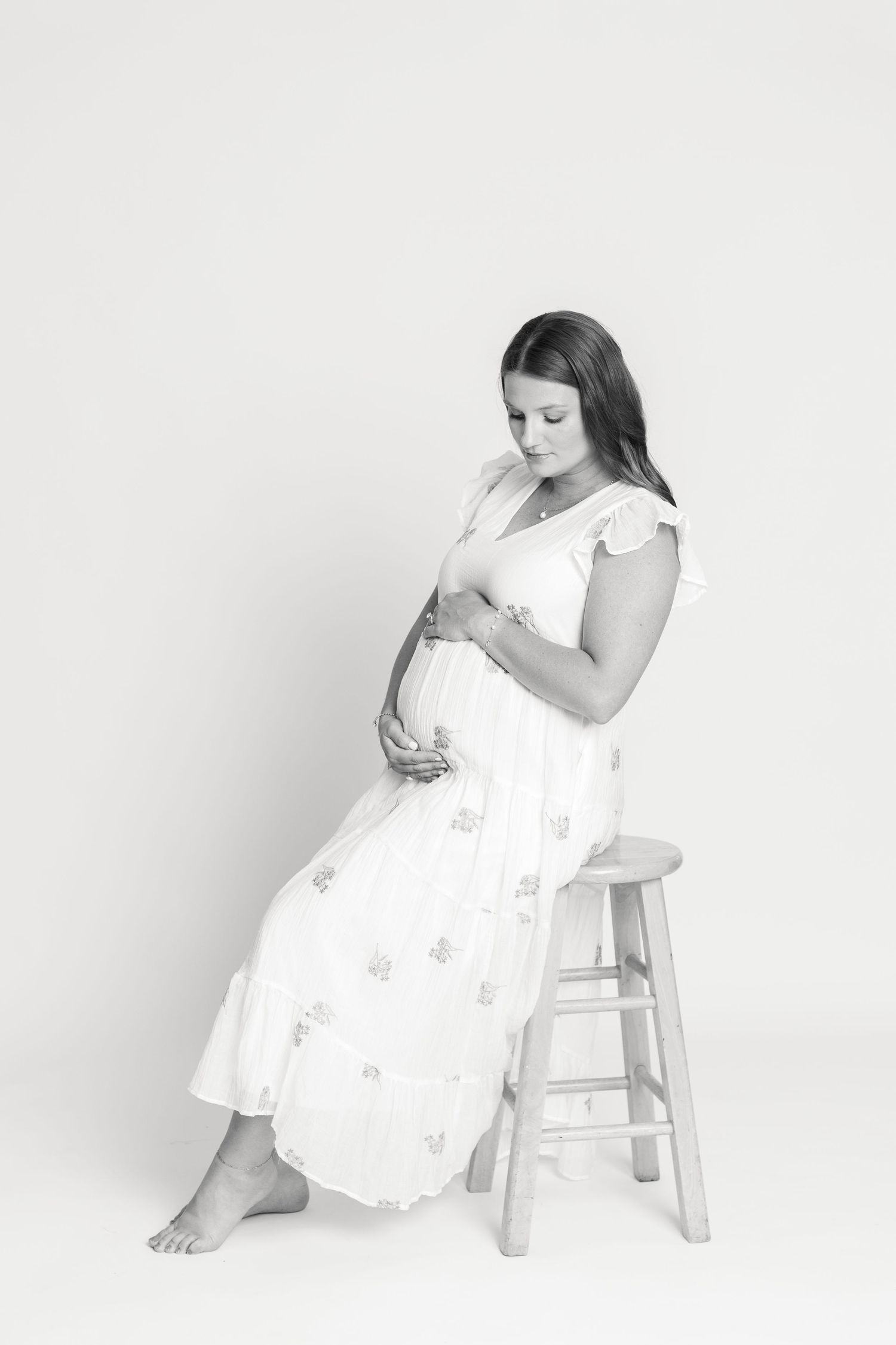 Black and white maternity photo of a woman in a white dress sitting on a stool, cradling her baby bump.