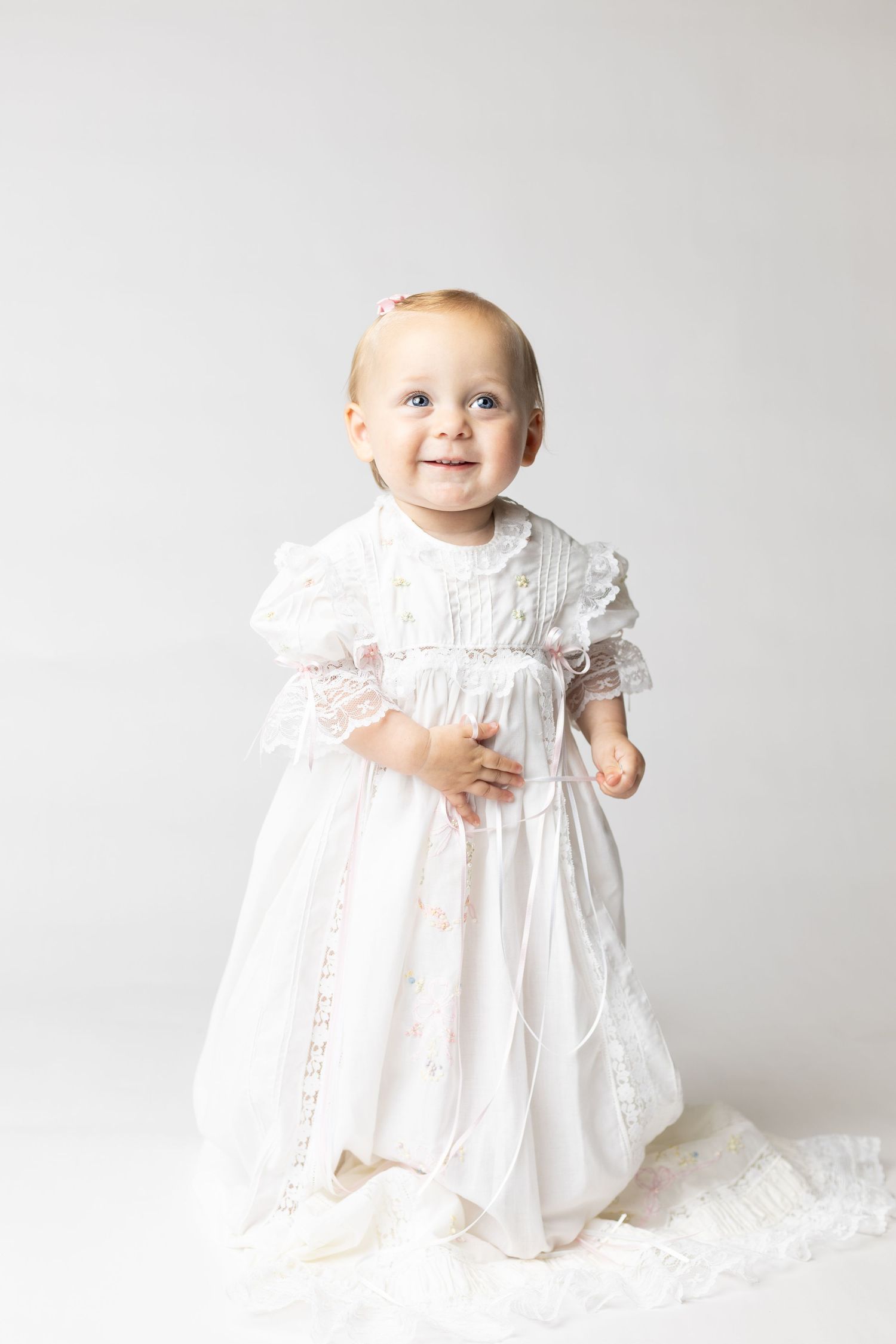 Smiling baby girl in white lace christening gown standing against light gray background.