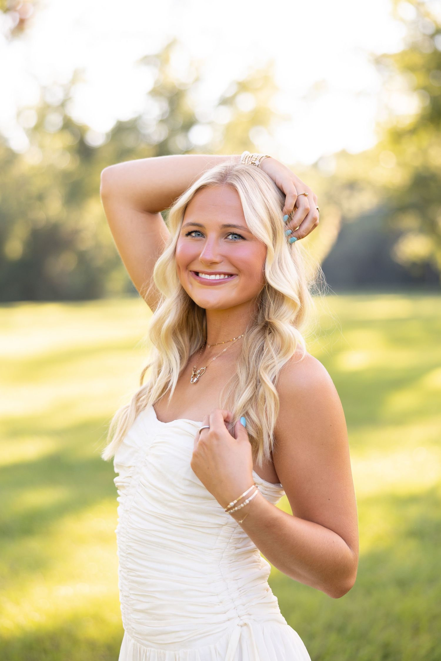 Smiling blonde woman in white strapless dress posing outdoors in sunlit green field with hand in hair.