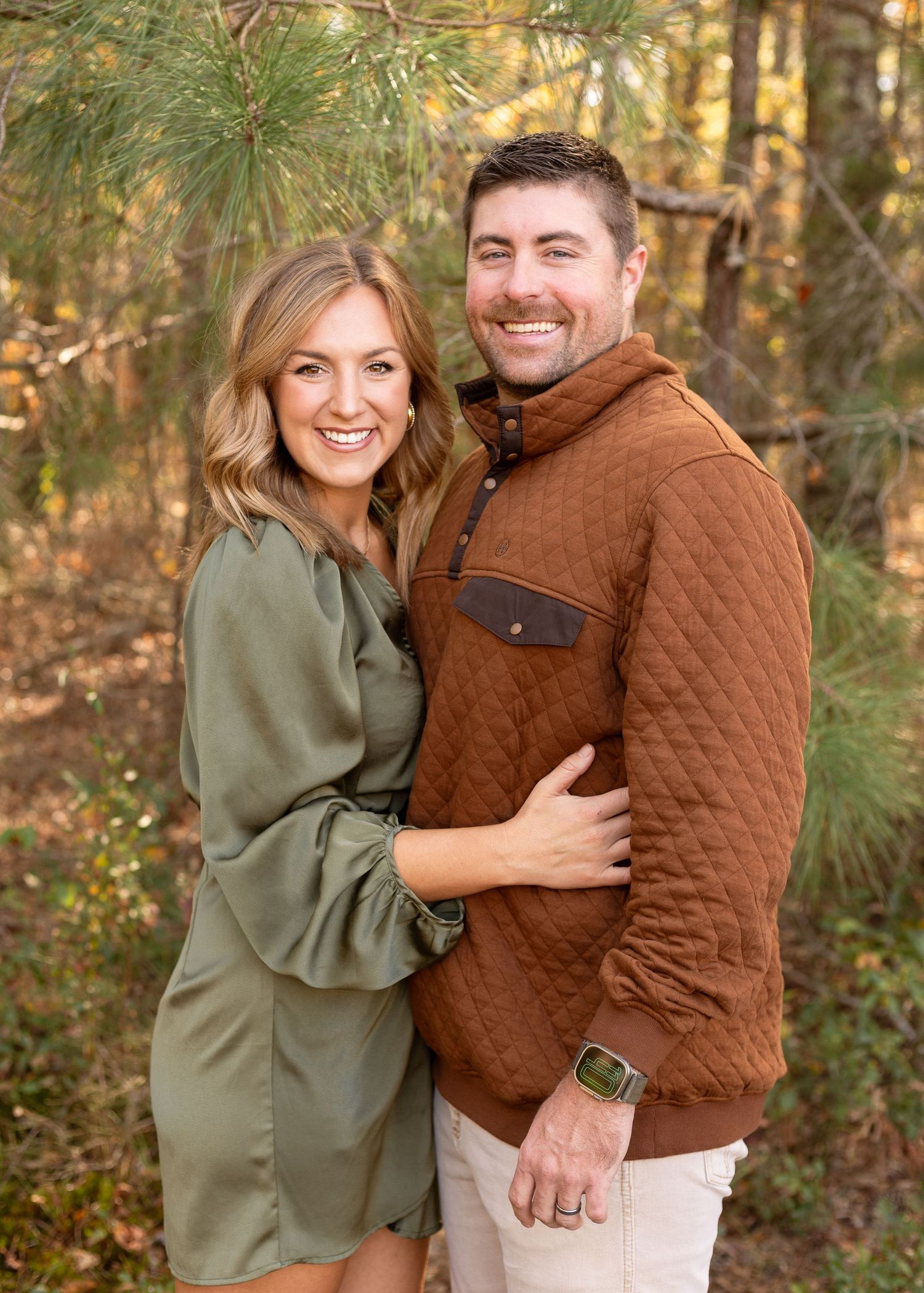 Smiling couple embracing outdoors in coordinated fall outfits among pine trees.