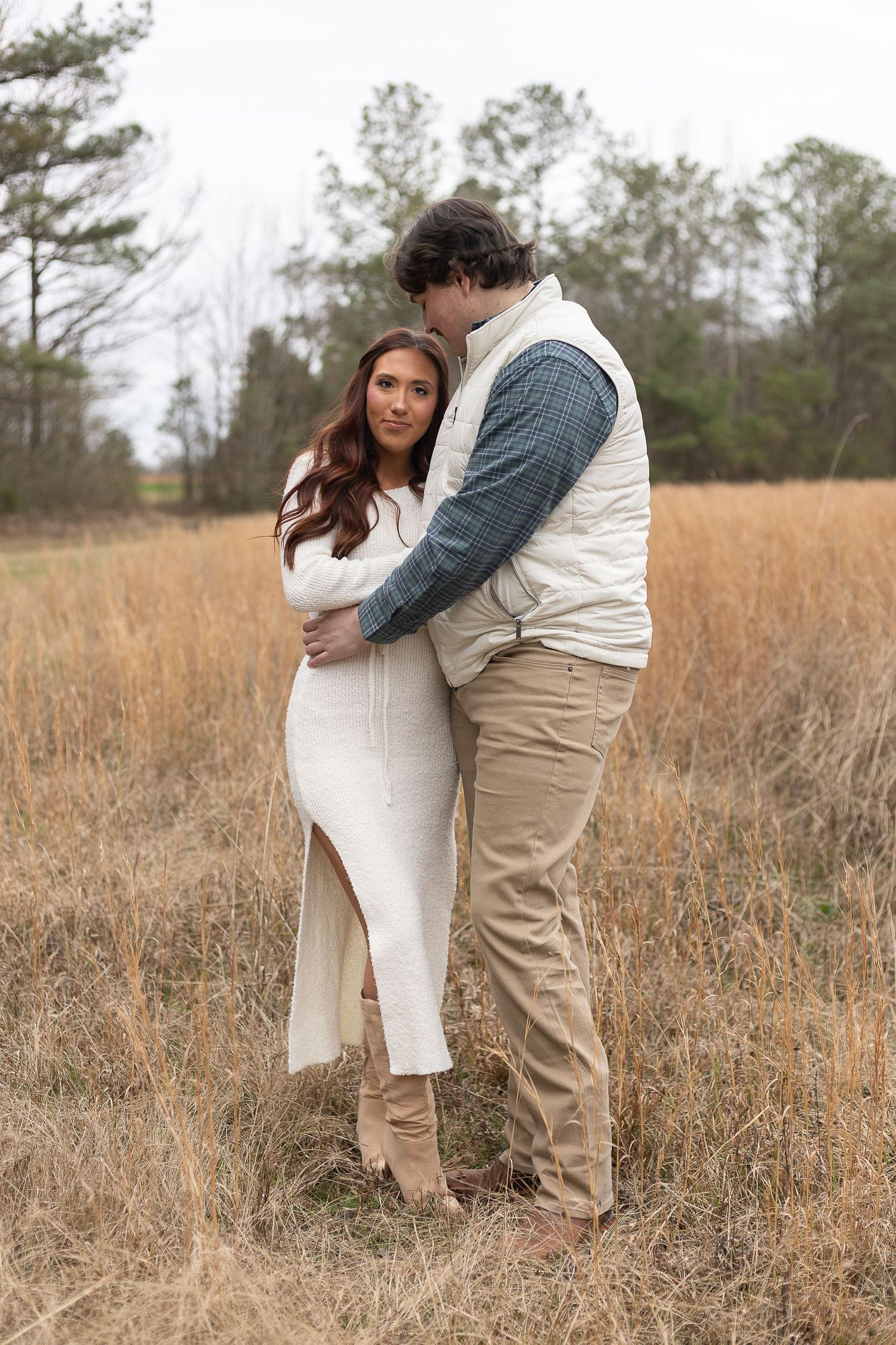 Couple embracing in golden field, woman in white dress, man in vest and khakis, trees in background.