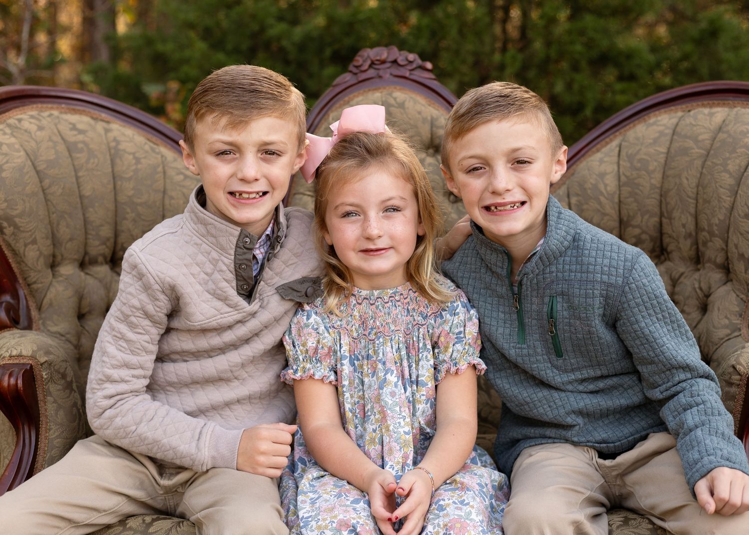 Three smiling children pose on a vintage sofa outdoors, two boys flanking a girl in a floral dress with a pink bow.