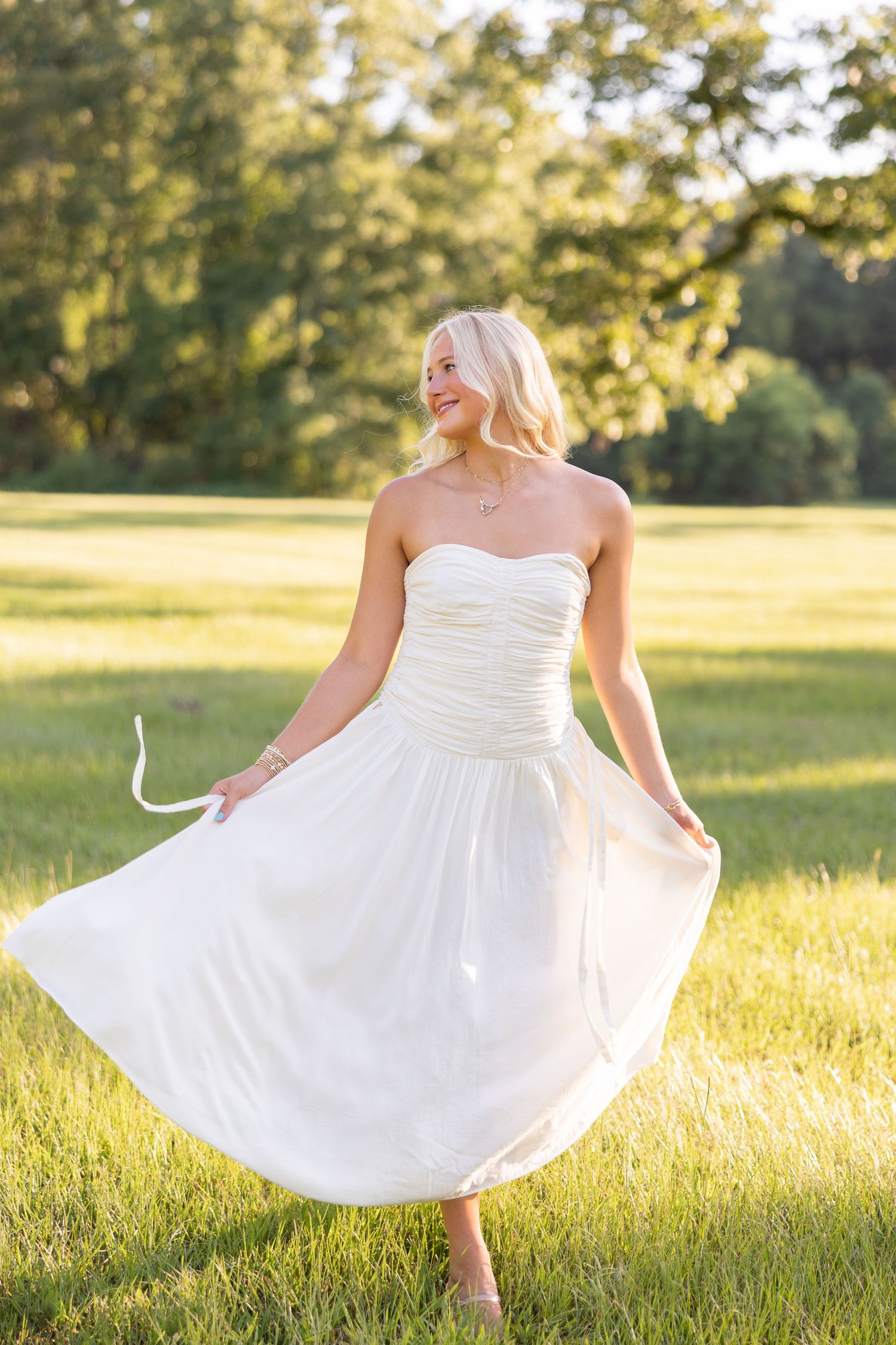 Blonde woman twirling in white strapless midi dress on green grass field with trees in background.