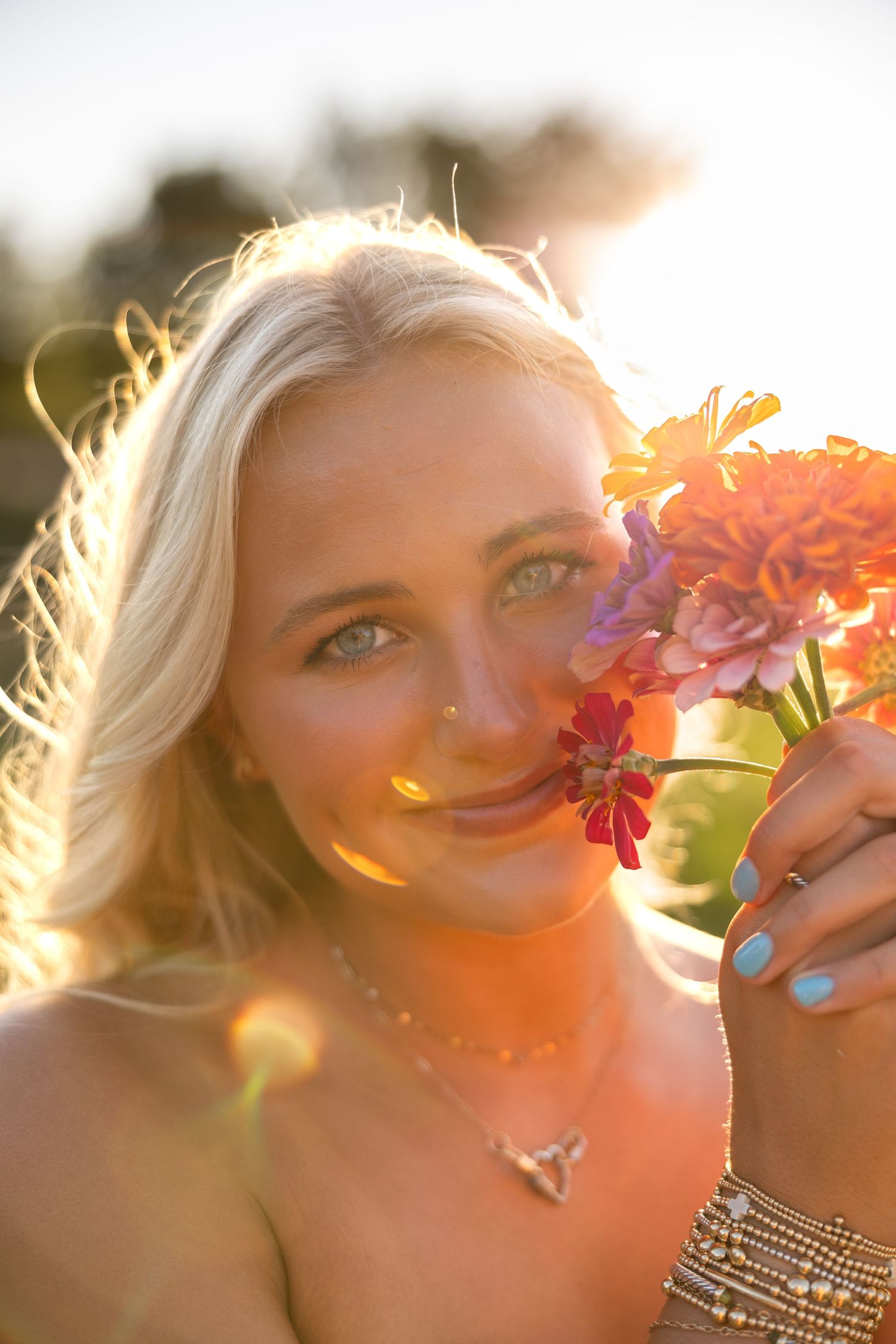 Blonde woman with blue eyes smelling colorful flowers in golden sunset light, wearing a necklace and blue nail polish.