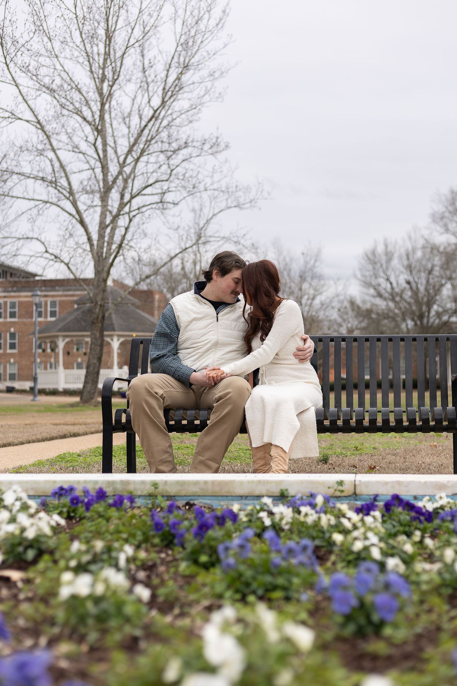Couple sharing a romantic moment on a park bench surrounded by blooming purple and white flowers on a cloudy day.