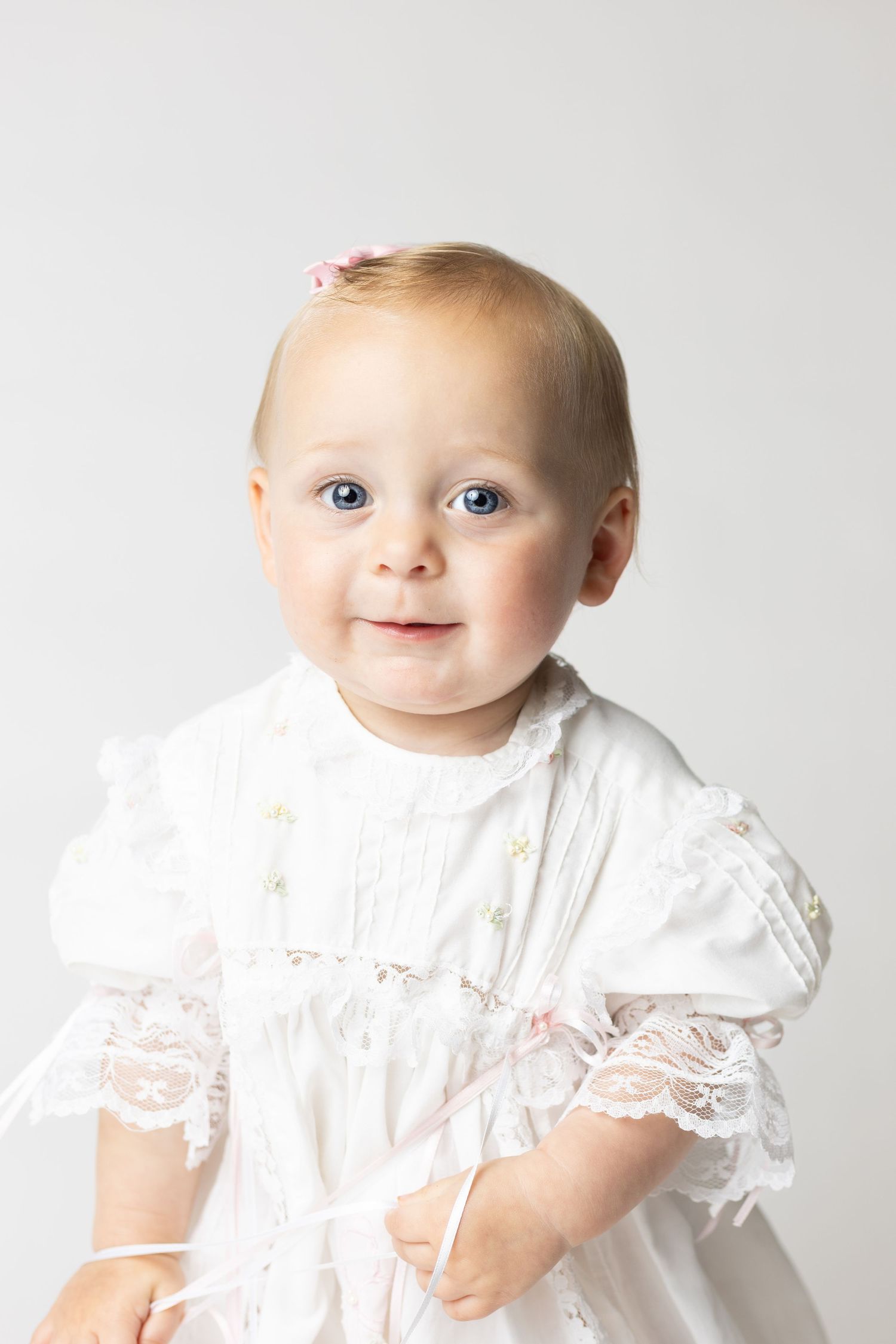 Adorable baby girl with blue eyes wearing a white lace christening dress and pink hair bow on a light background.