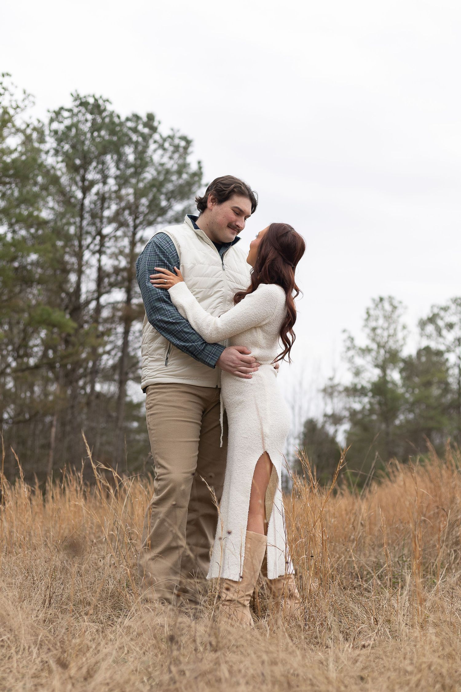 Couple embracing in a golden field, man in white vest and khakis, woman in white dress and boots, pine trees behind.
