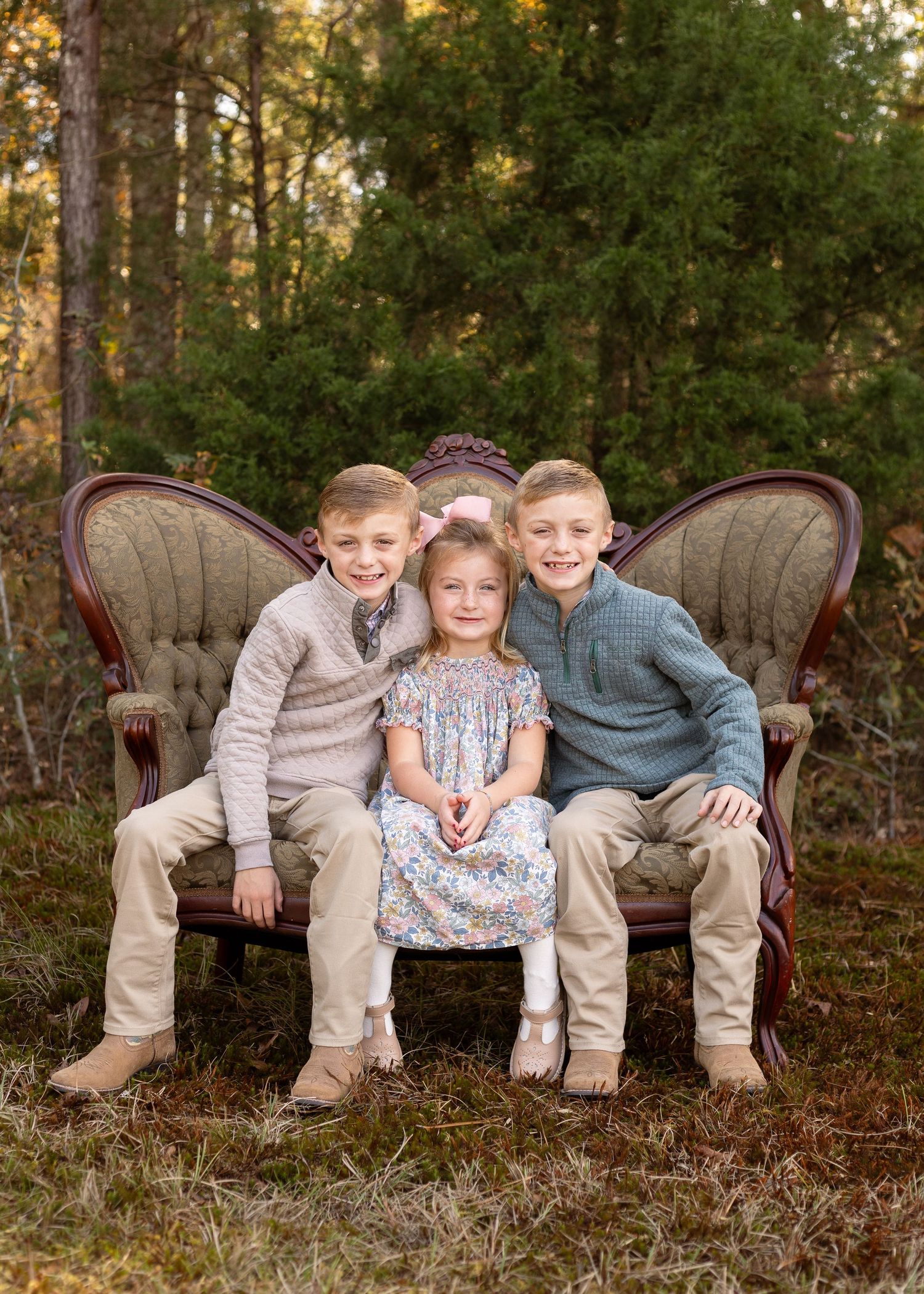 Three children pose on a vintage Victorian settee outdoors, two boys flanking a girl in a floral dress.