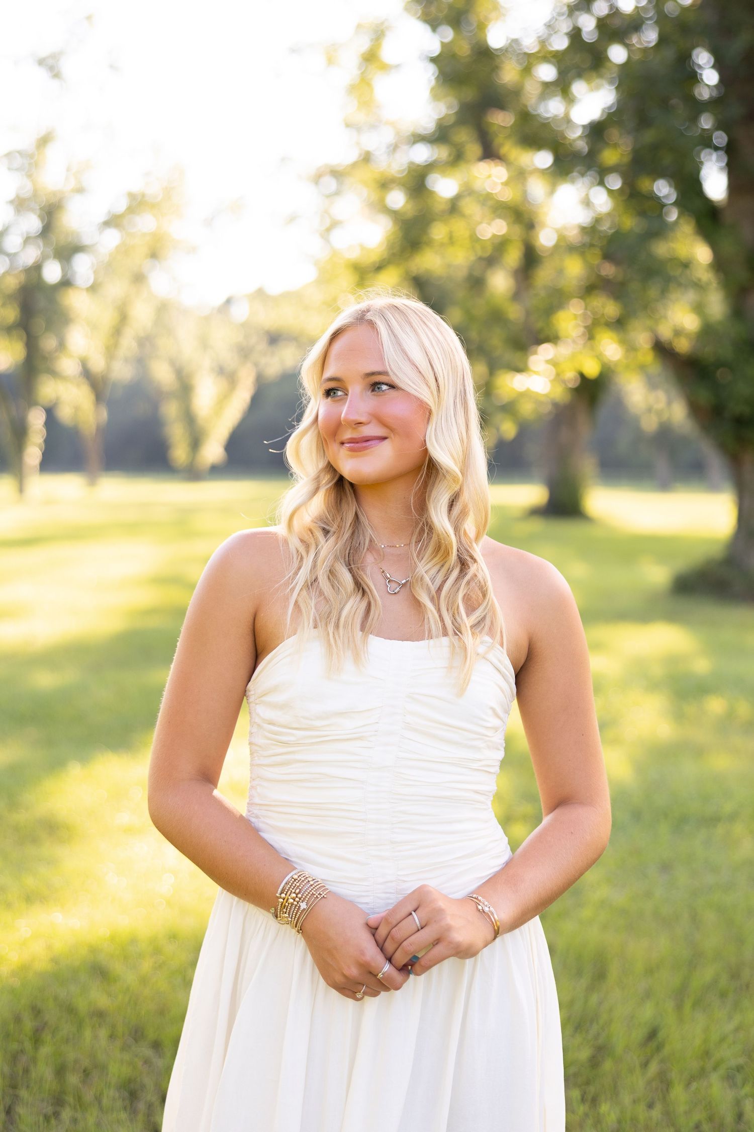 Young blonde woman in white strapless dress smiling outdoors in sunlit green park setting.
