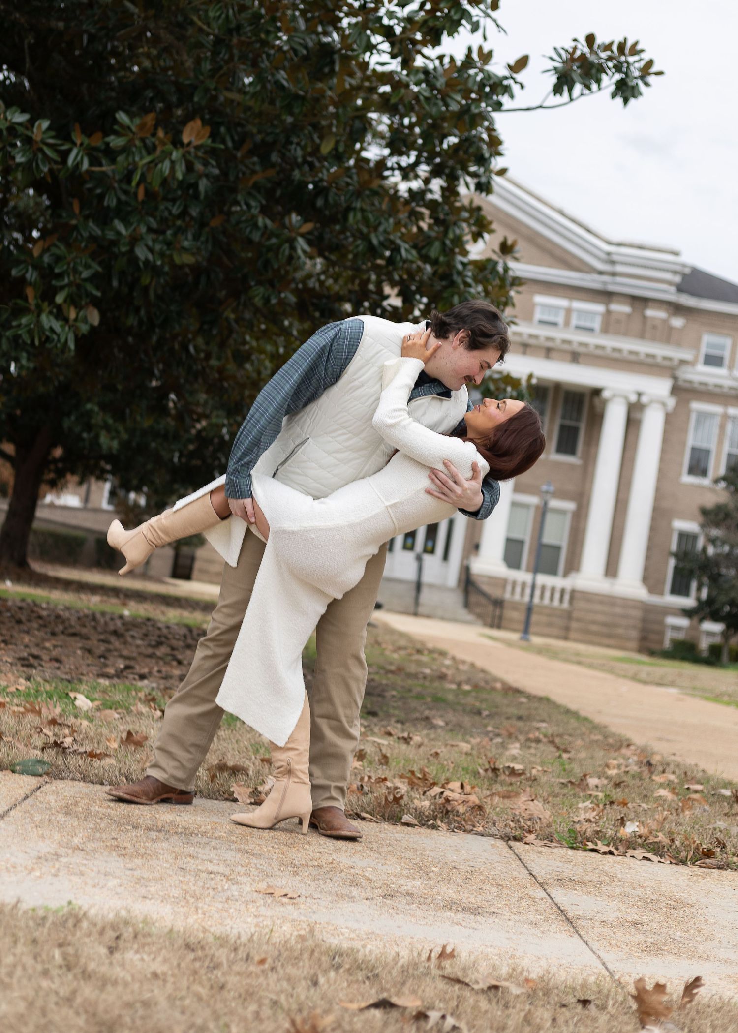 Couple sharing a romantic dip kiss on a sidewalk outside a historic brick building surrounded by trees.