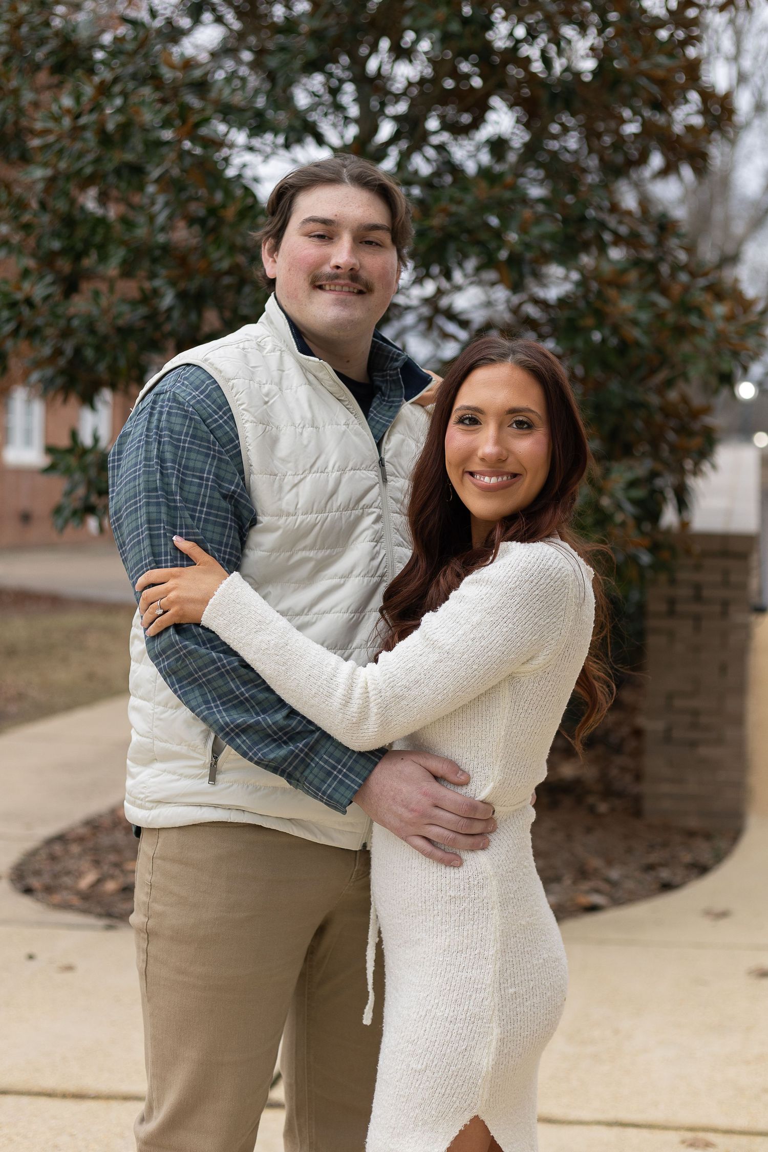 Smiling couple posing together outdoors, woman in white dress and man in plaid shirt and white vest hugging near trees.