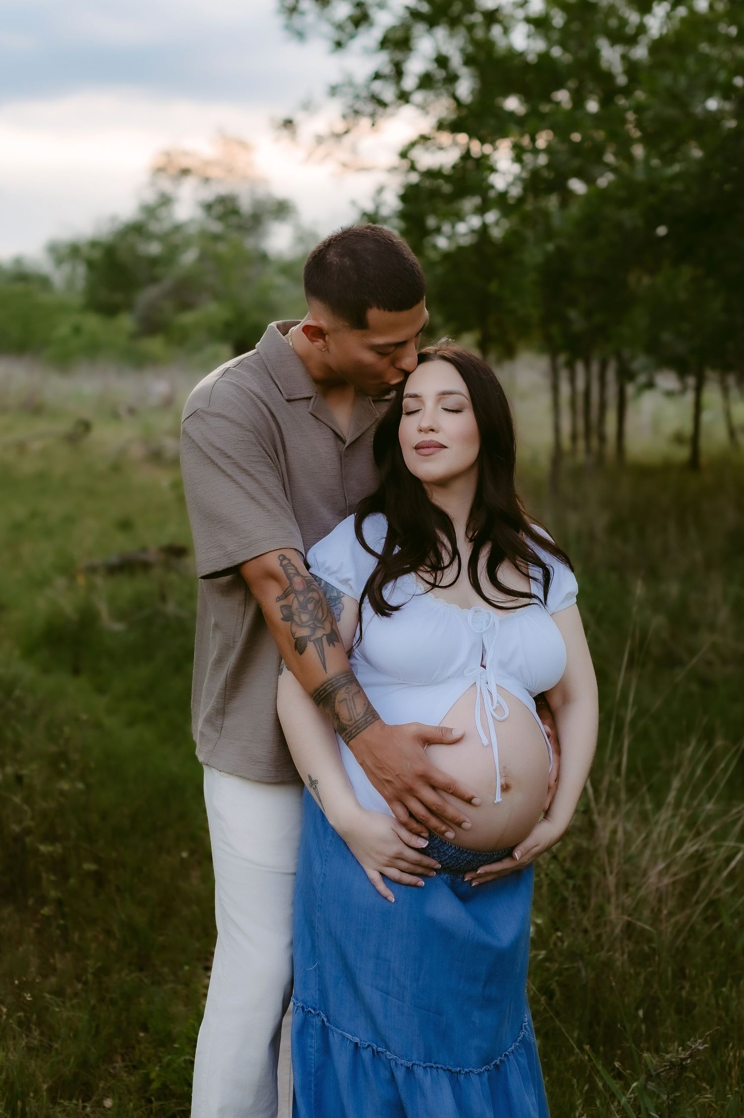 An expectant couple embraces in a serene outdoor setting with tall grass and trees at sunset.