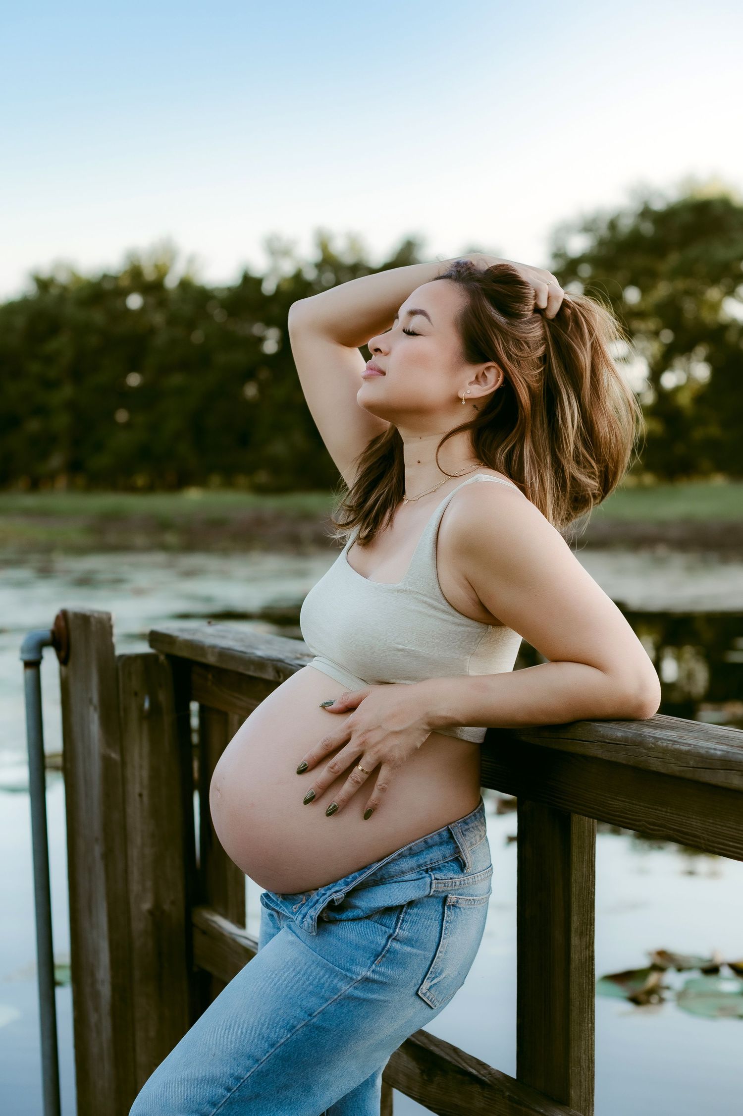 A pregnant woman in jeans and crop top poses against a wooden dock railing during sunset by a lake.