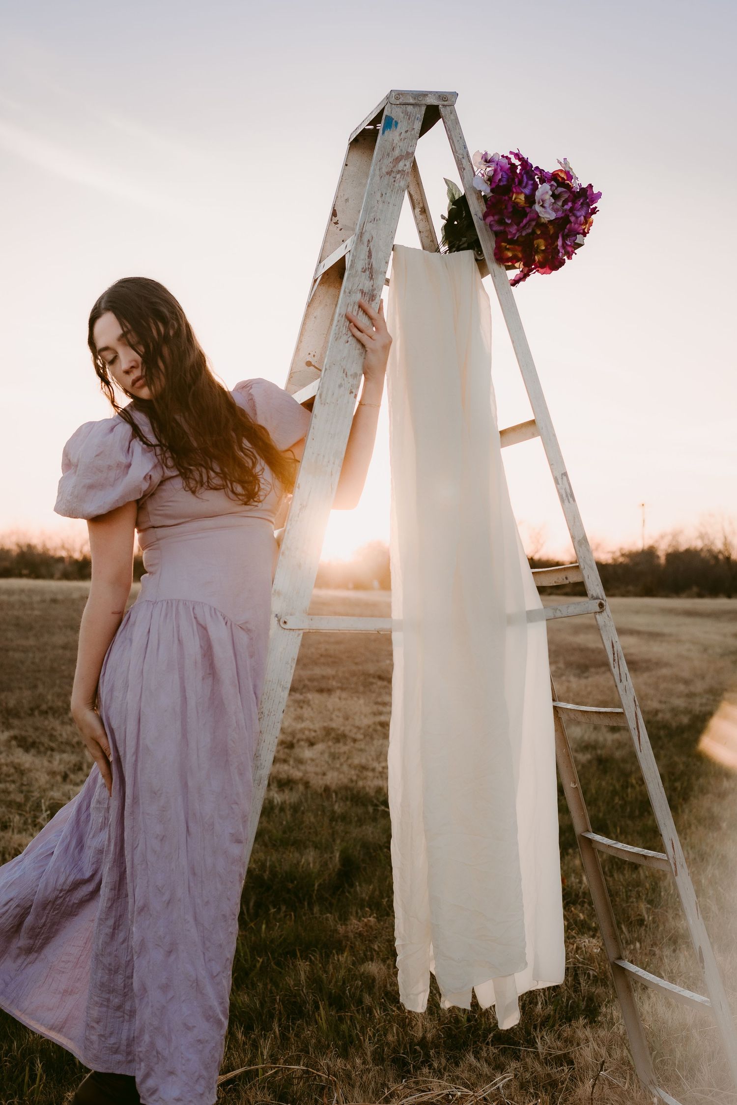 A vintage-style scene with a ladder, hanging dress and flowers at sunset in a field.