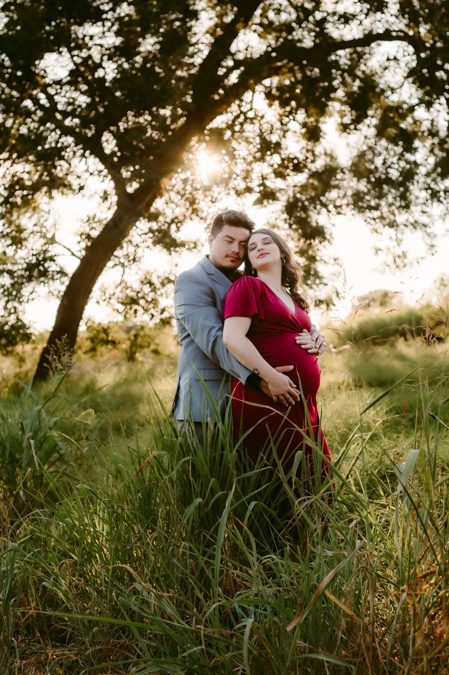 A romantic couple embraces in tall grass at sunset, silhouetted by golden sunlight filtering through tree branches.