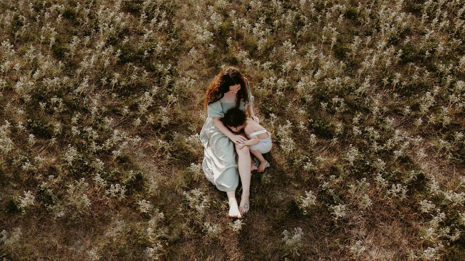 An aerial view shows someone in a white dress lying amidst a field of small white wildflowers.