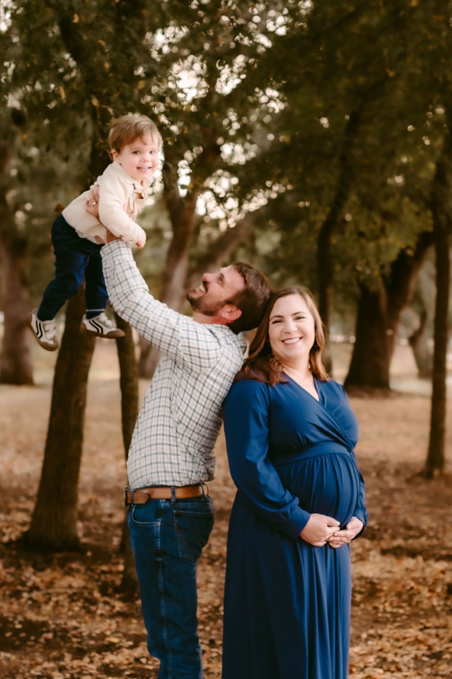 A family poses together in a wooded park during autumn with fallen leaves on the ground.