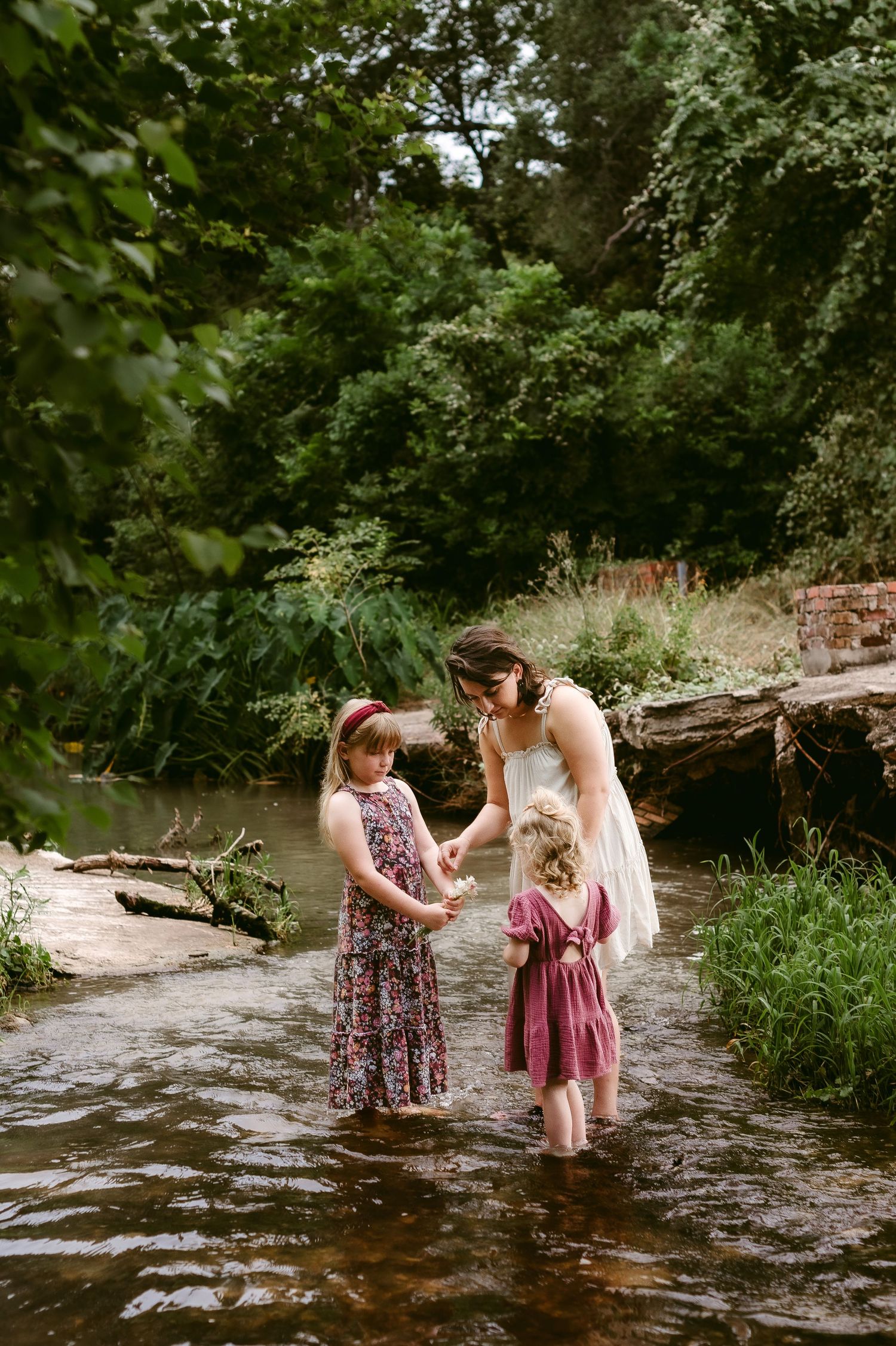 Three people in flowy dresses stand together in a shallow stream surrounded by lush green trees on a summer day.