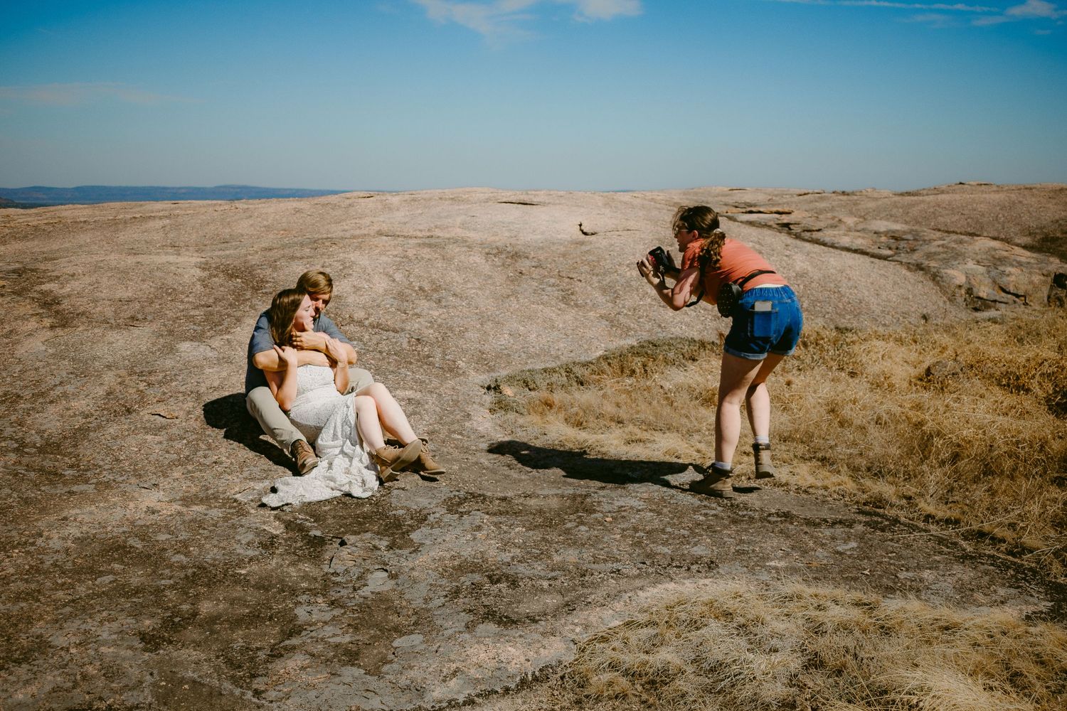 A photographer in shorts captures a scenic photoshoot on a rugged coastal cliff against a bright blue sky.