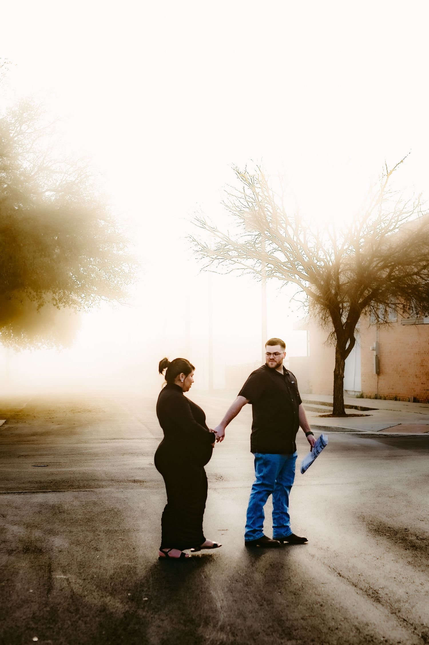Two people stand together on a misty road at dawn with bare trees creating a moody silhouette in the background.