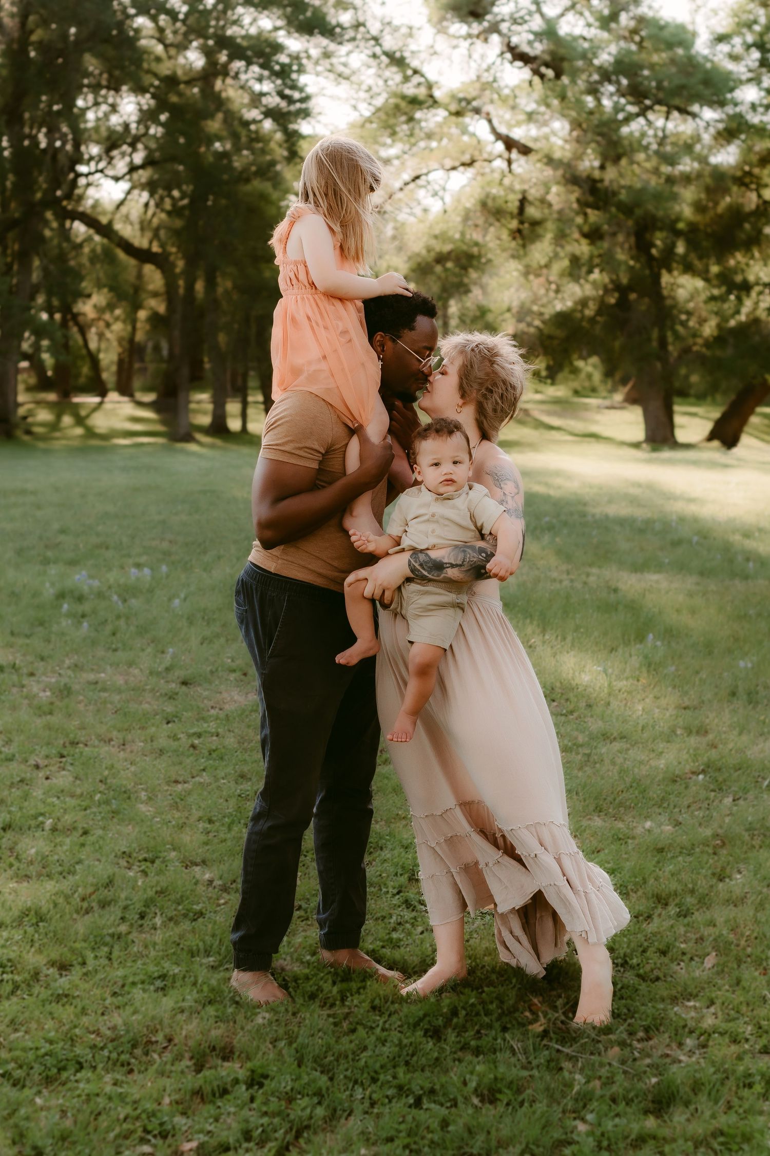 A family embraces together in a sunlit park with soft lighting filtering through the trees on a warm afternoon.