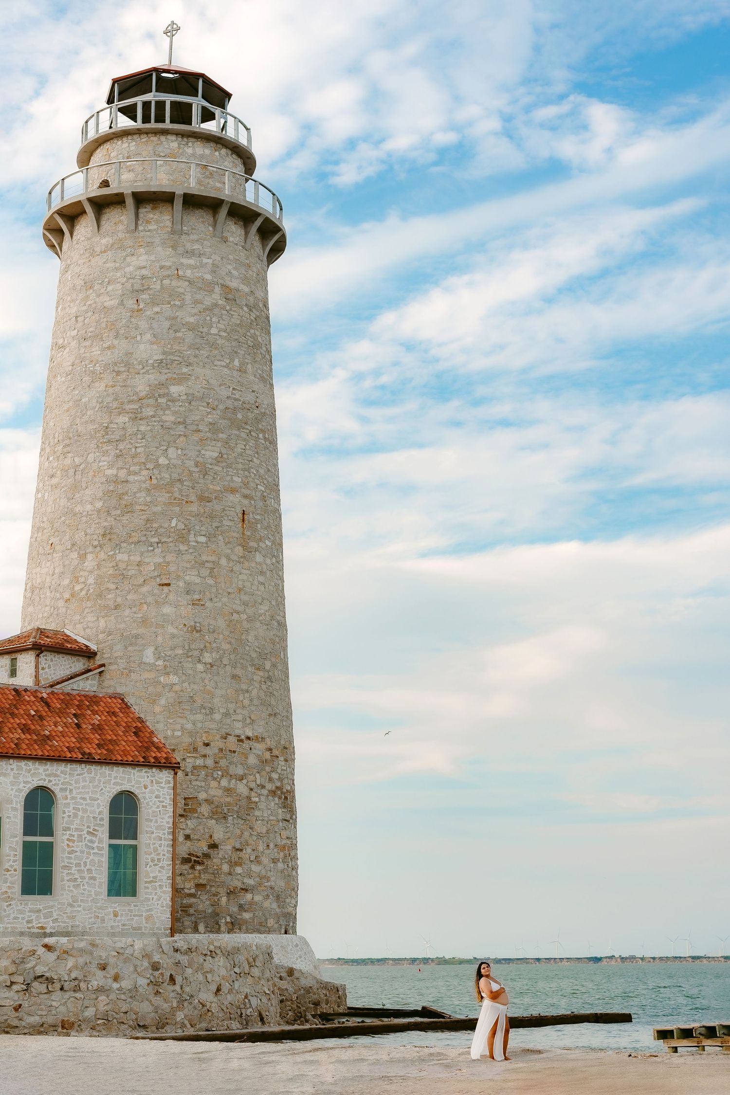 A tall white lighthouse stands against a bright blue sky on a rocky shoreline next to calm waters, with orange tiled building.