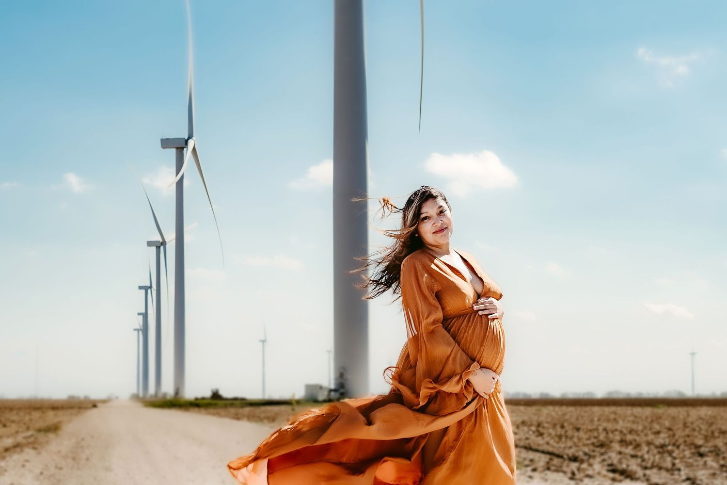 A flowing orange dress contrasts against white wind turbines on a rural dirt road under blue sky.