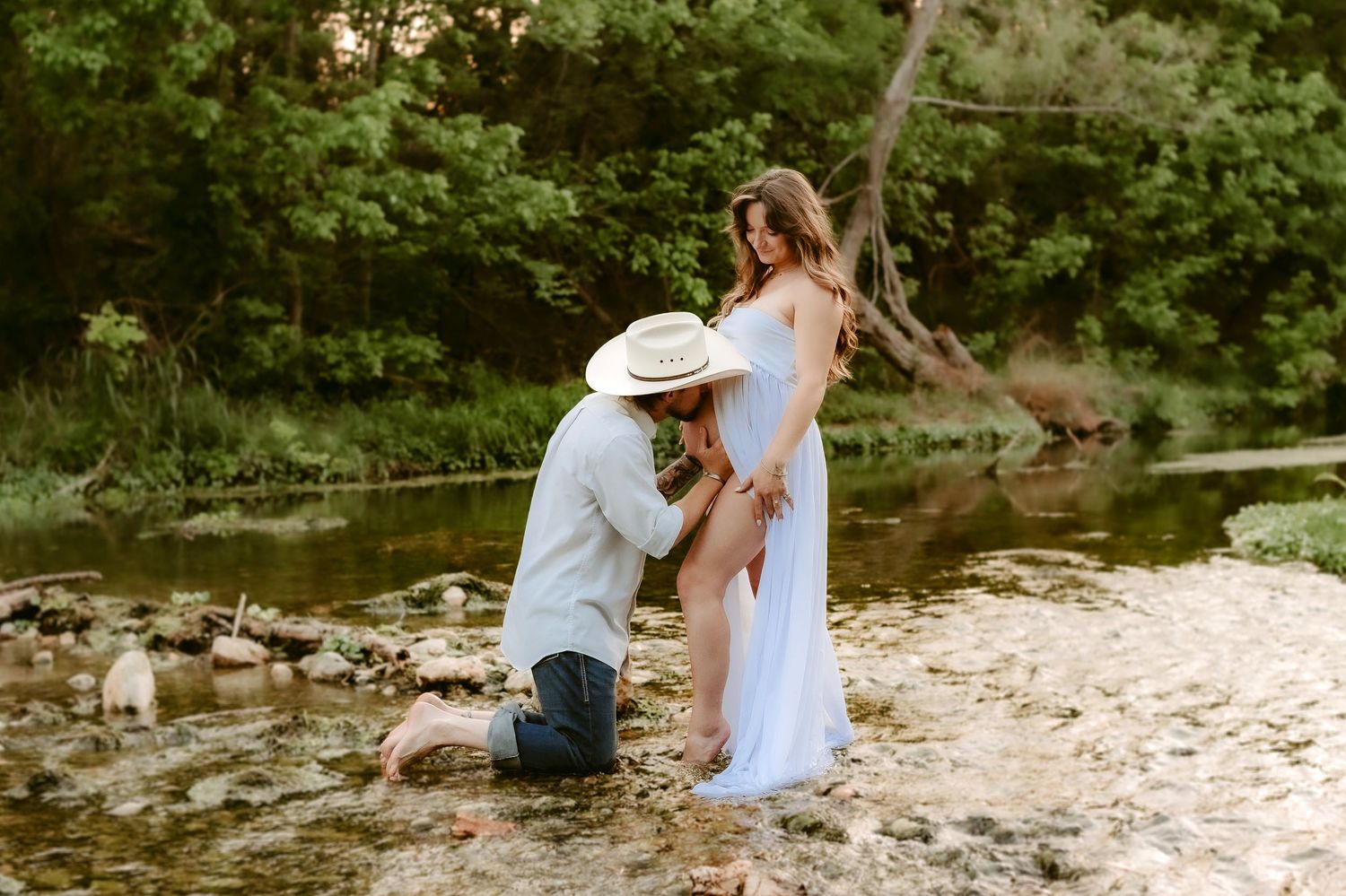 A couple shares an intimate moment on the rocky shores of a peaceful creek in a lush forest setting.