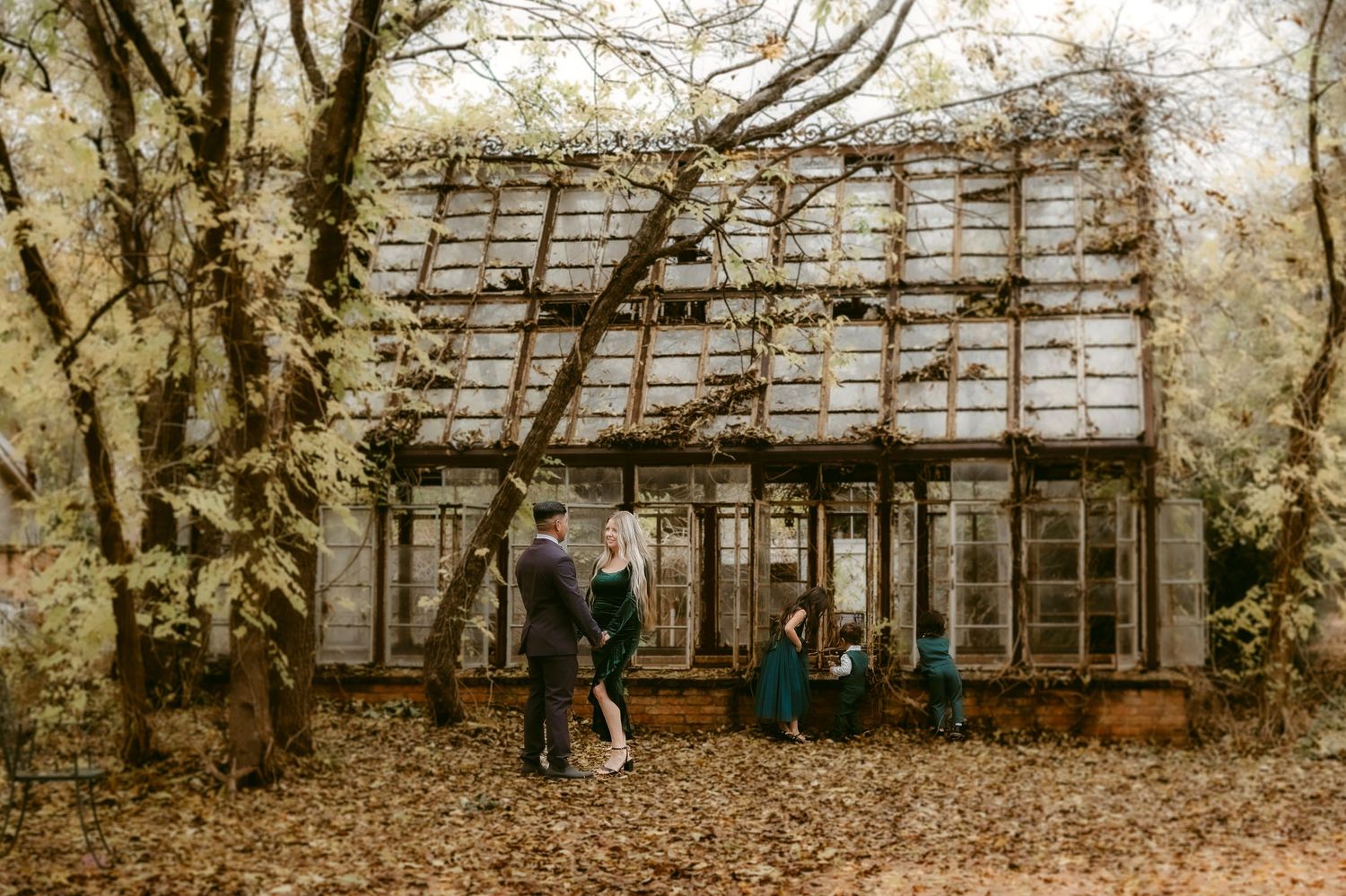 Couple stands outside a rustic greenhouse or glasshouse structure in an autumn woodland setting.