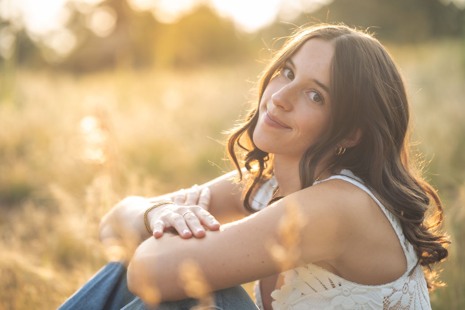 Young woman with dark hair sitting in a sunlit golden field, smiling softly with arms crossed over knees.