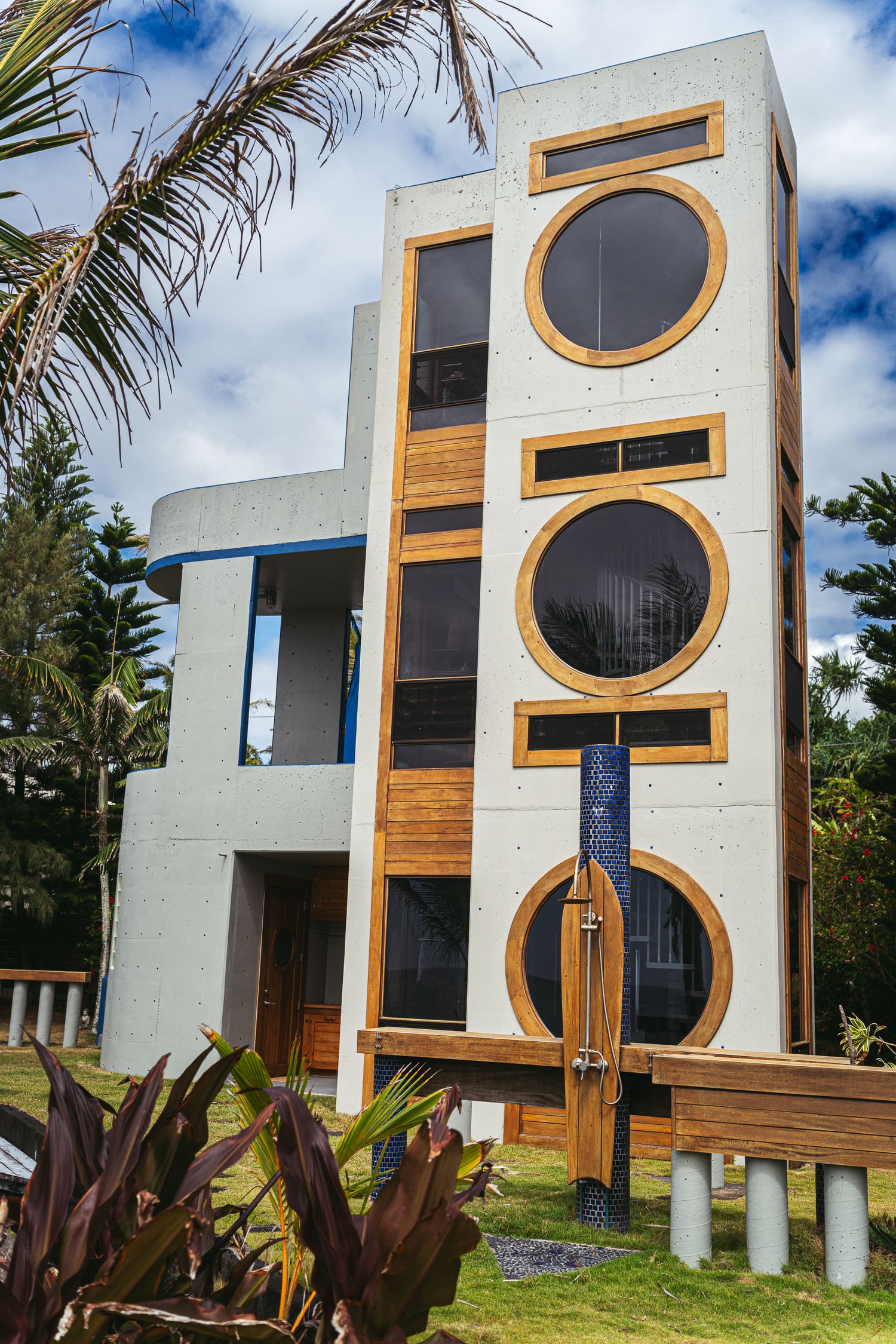 Modern multi-story concrete and wood building with circular windows, surrounded by tropical plants under a blue sky.