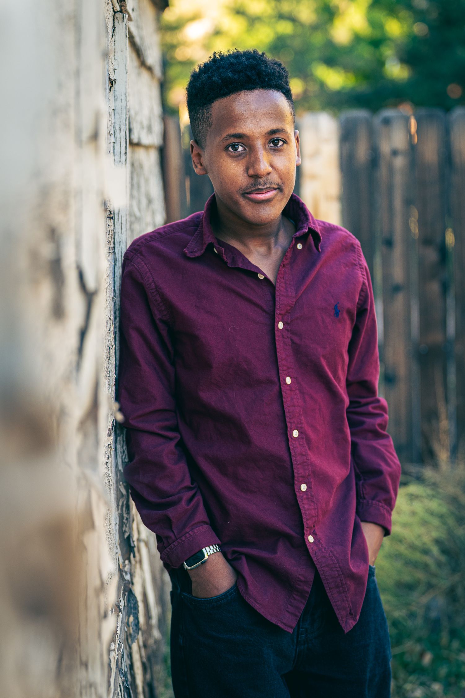 Young man in burgundy Ralph Lauren shirt leaning against a rustic wall outdoors, hands in pockets.