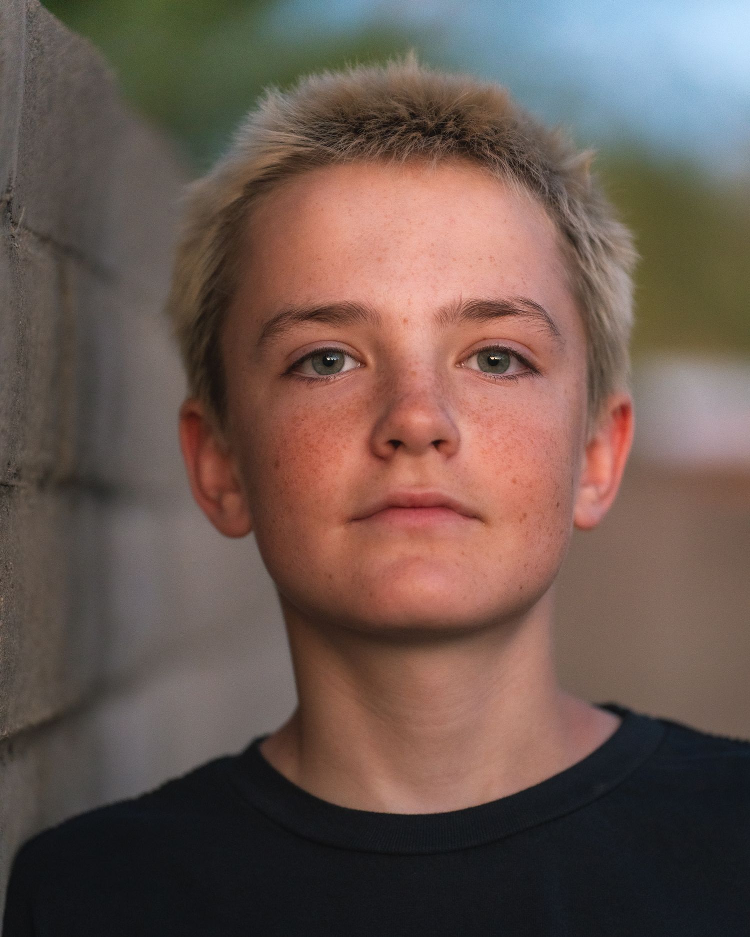 Close-up portrait of a young person with short blonde hair, freckles, and green eyes wearing a black shirt.
