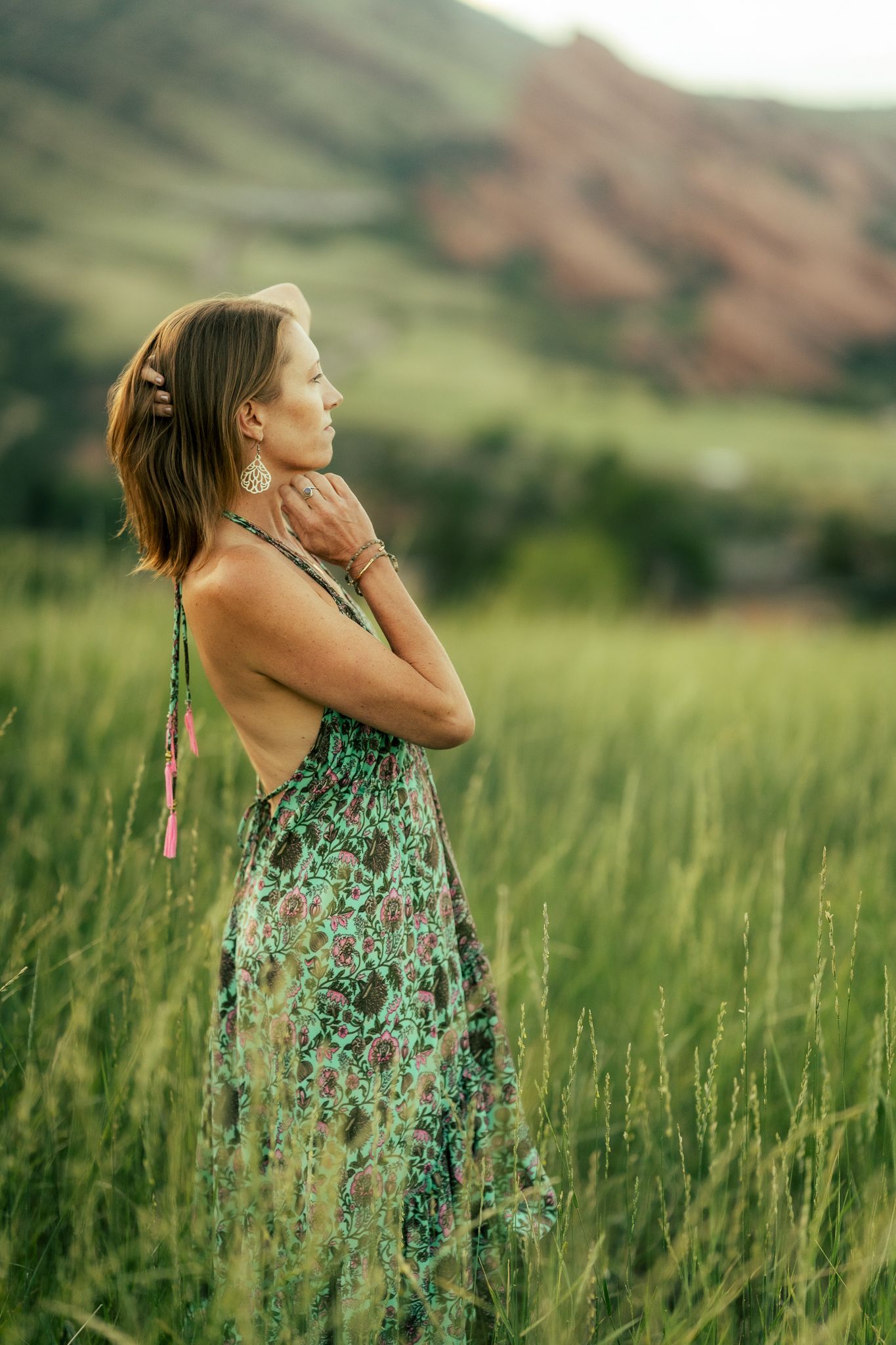 A person in a floral print sundress stands peacefully in a tall grass meadow with red rocks visible in the background.