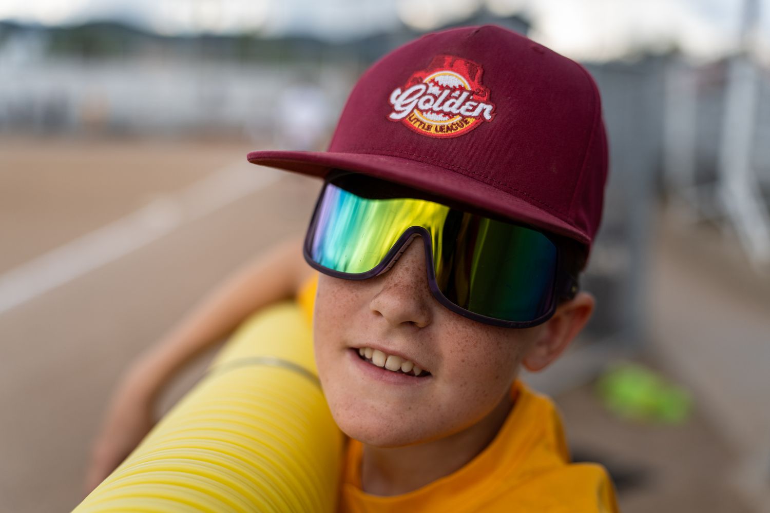 Young baseball fan wearing a maroon cap and colorful sunglasses at a sports field.