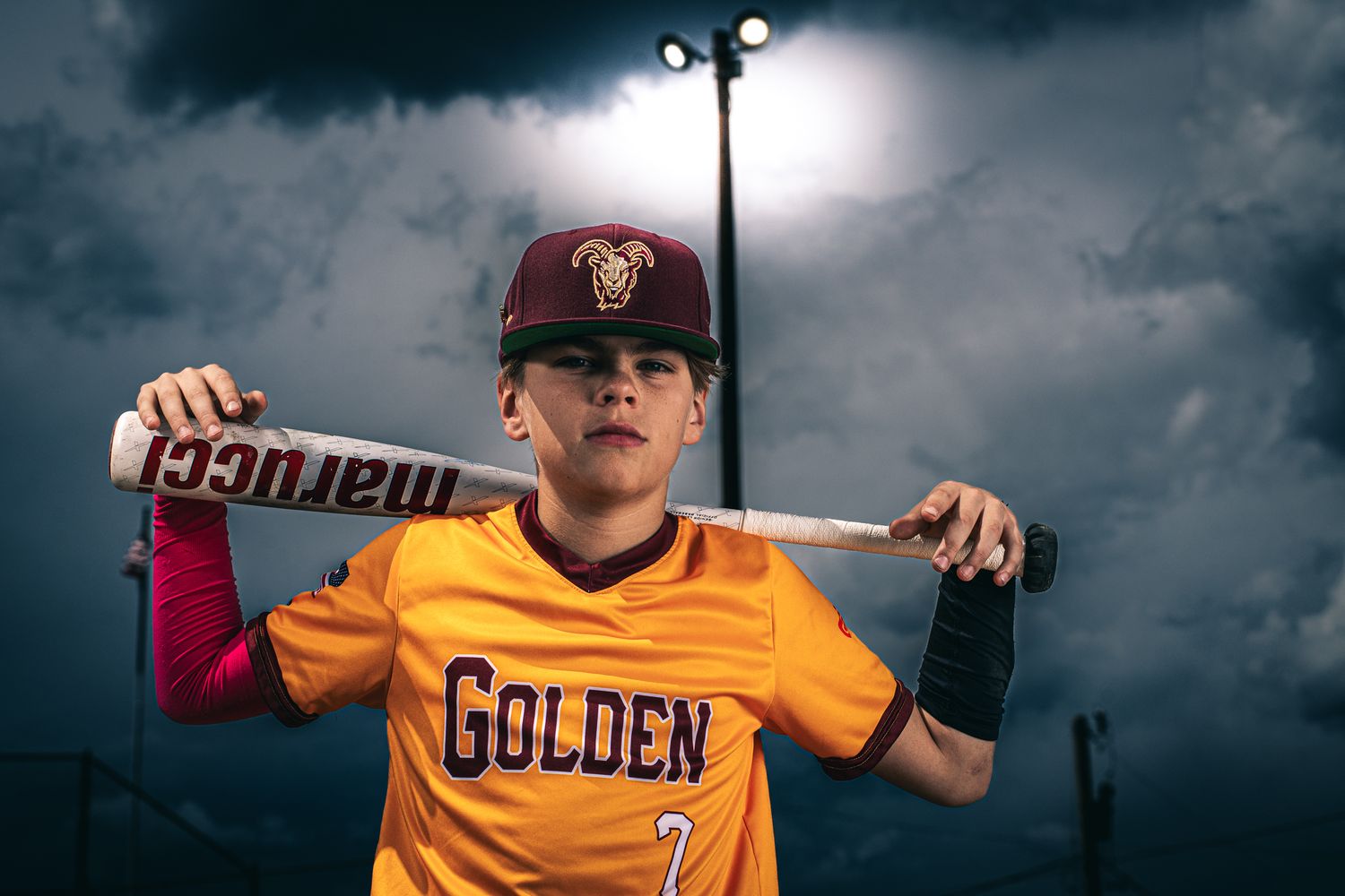 Youth baseball players in golden jerseys pose under dramatic storm clouds with stadium lighting overhead.