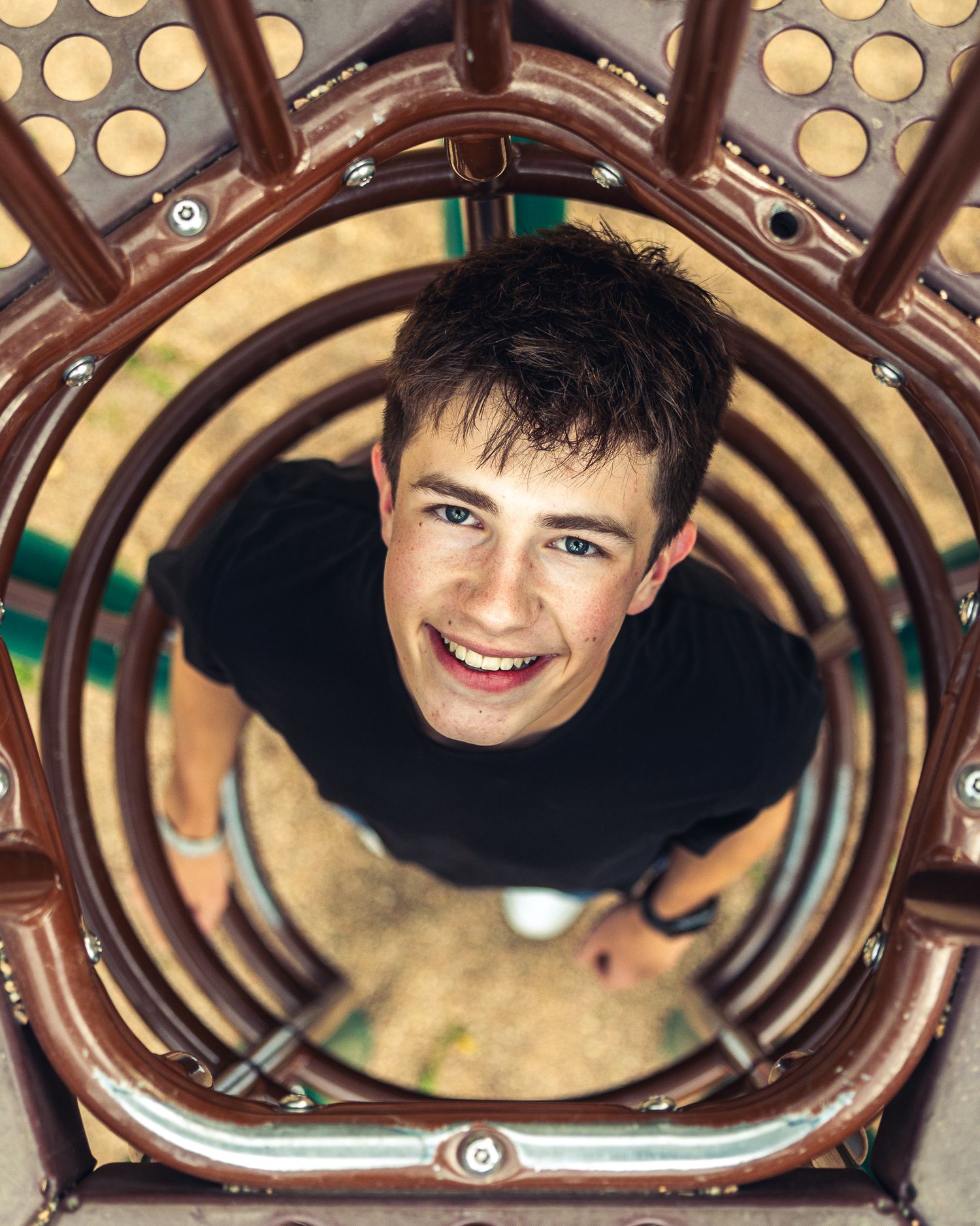 Smiling young man photographed from above through a circular tunnel on a playground structure.