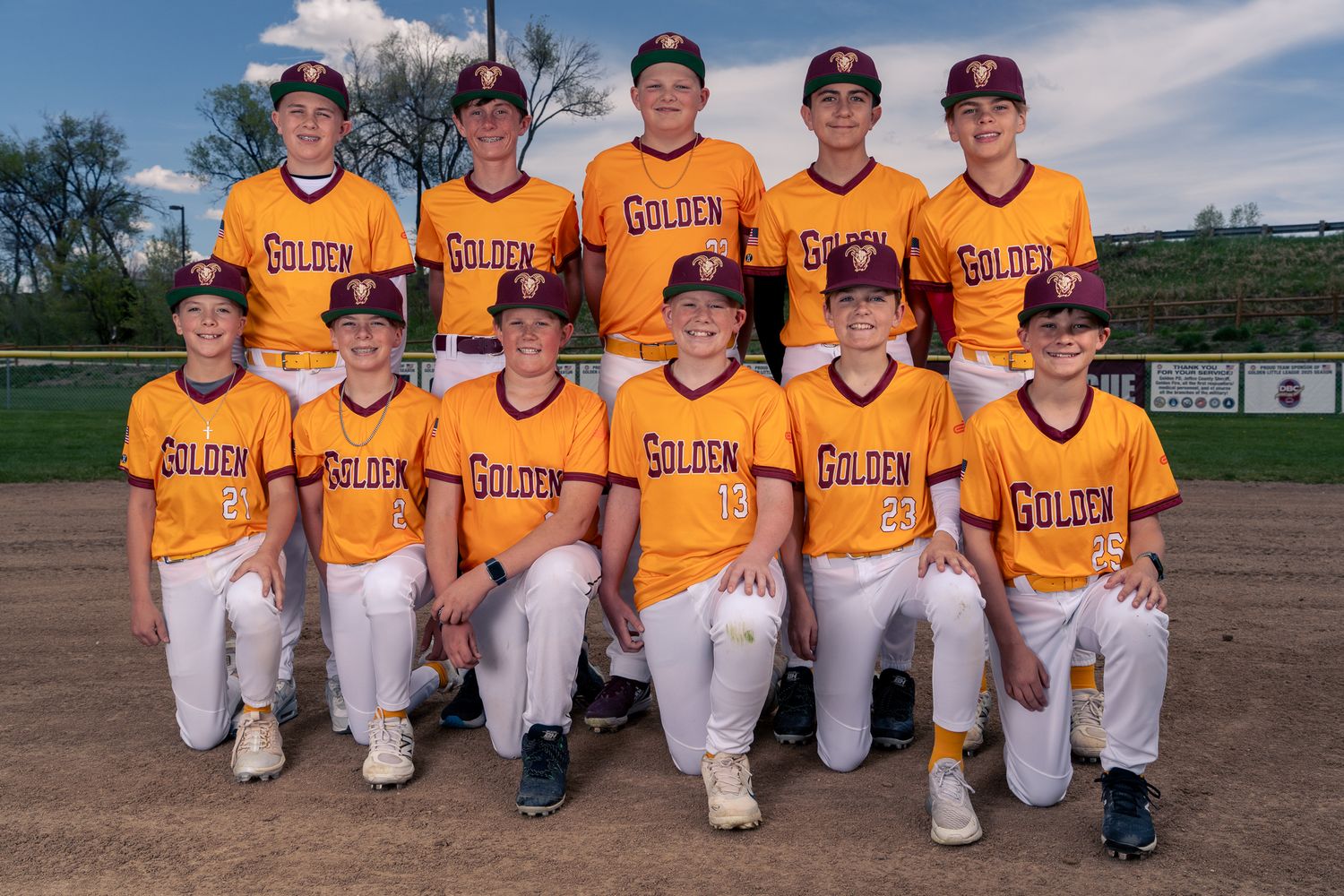 Youth baseball players in yellow jerseys kneel in a row on a baseball field during a team photo session.