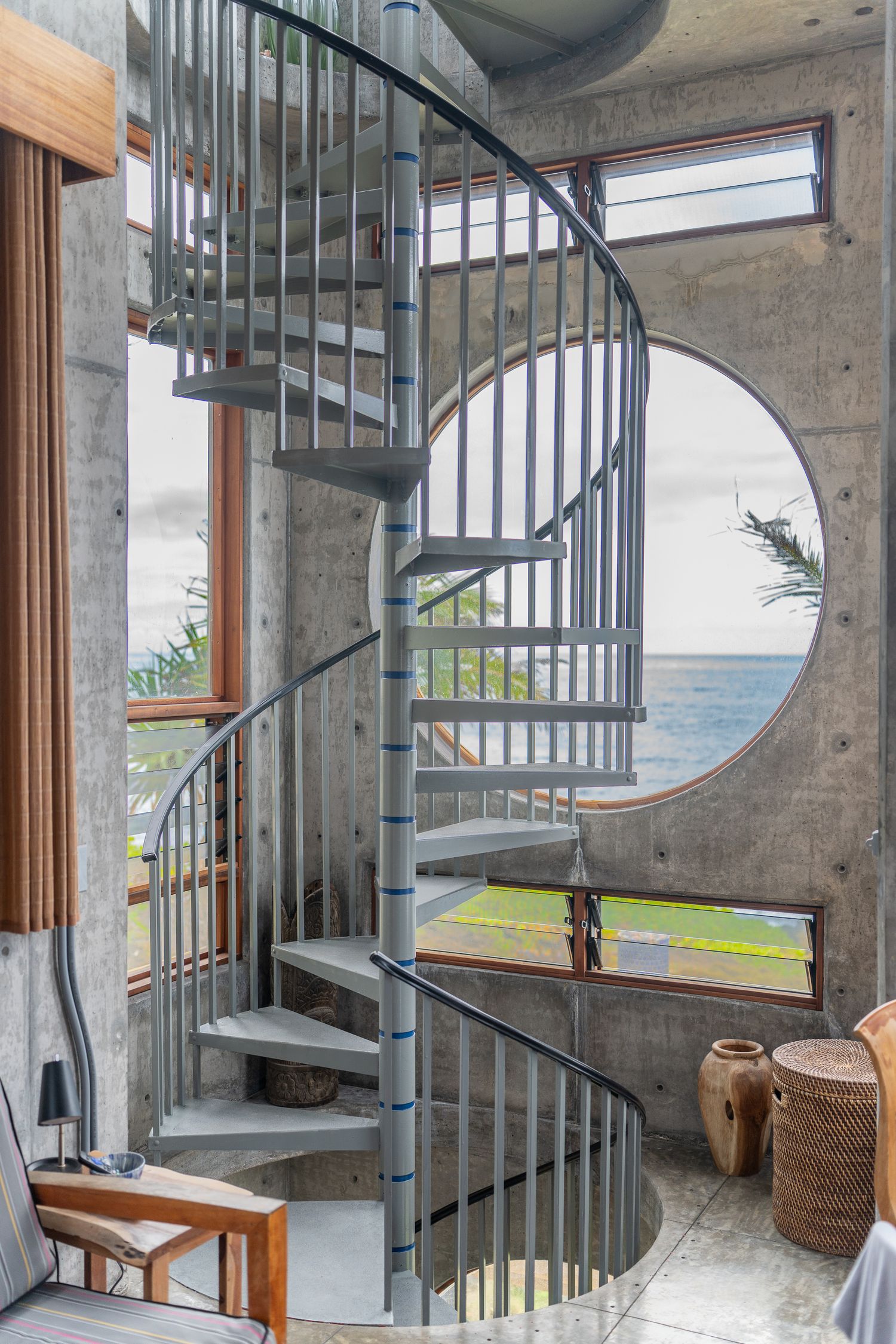 Spiral metal staircase in a concrete interior with a circular ocean-view window and wooden accents.