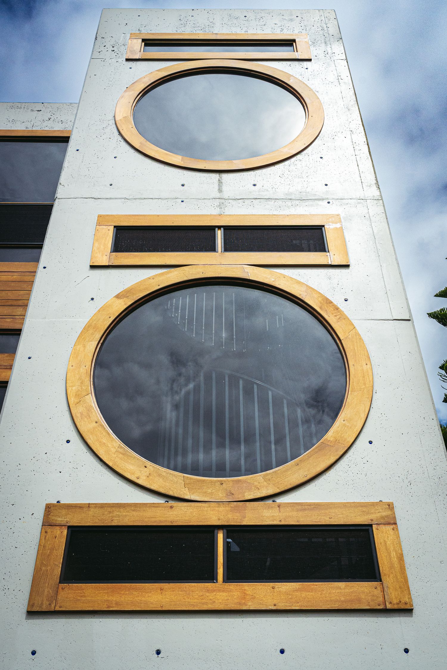 Modern concrete building facade featuring circular and rectangular wooden-framed windows against a cloudy sky.