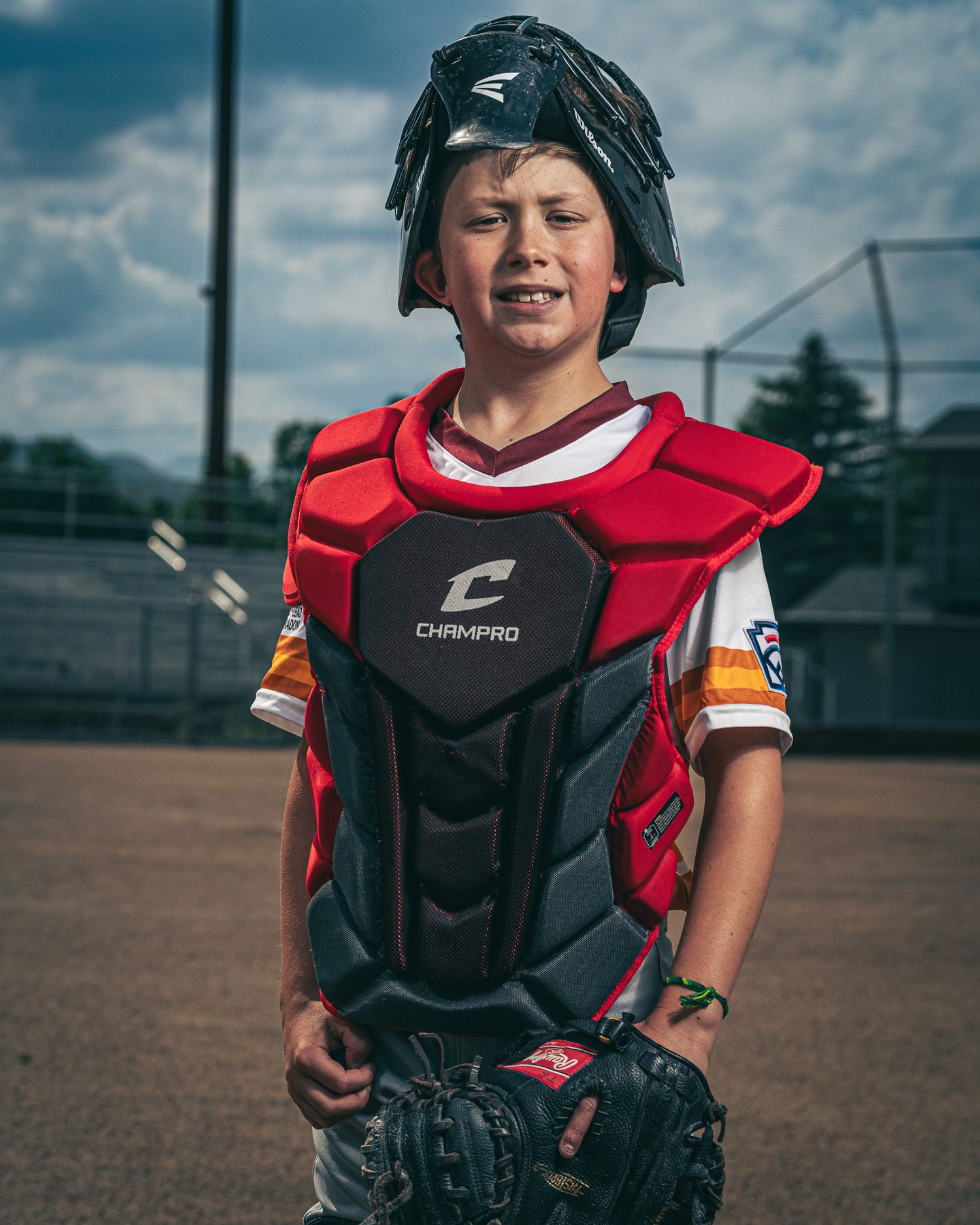 Young baseball catcher in protective gear kneels on dusty field during practice on a cloudy day.